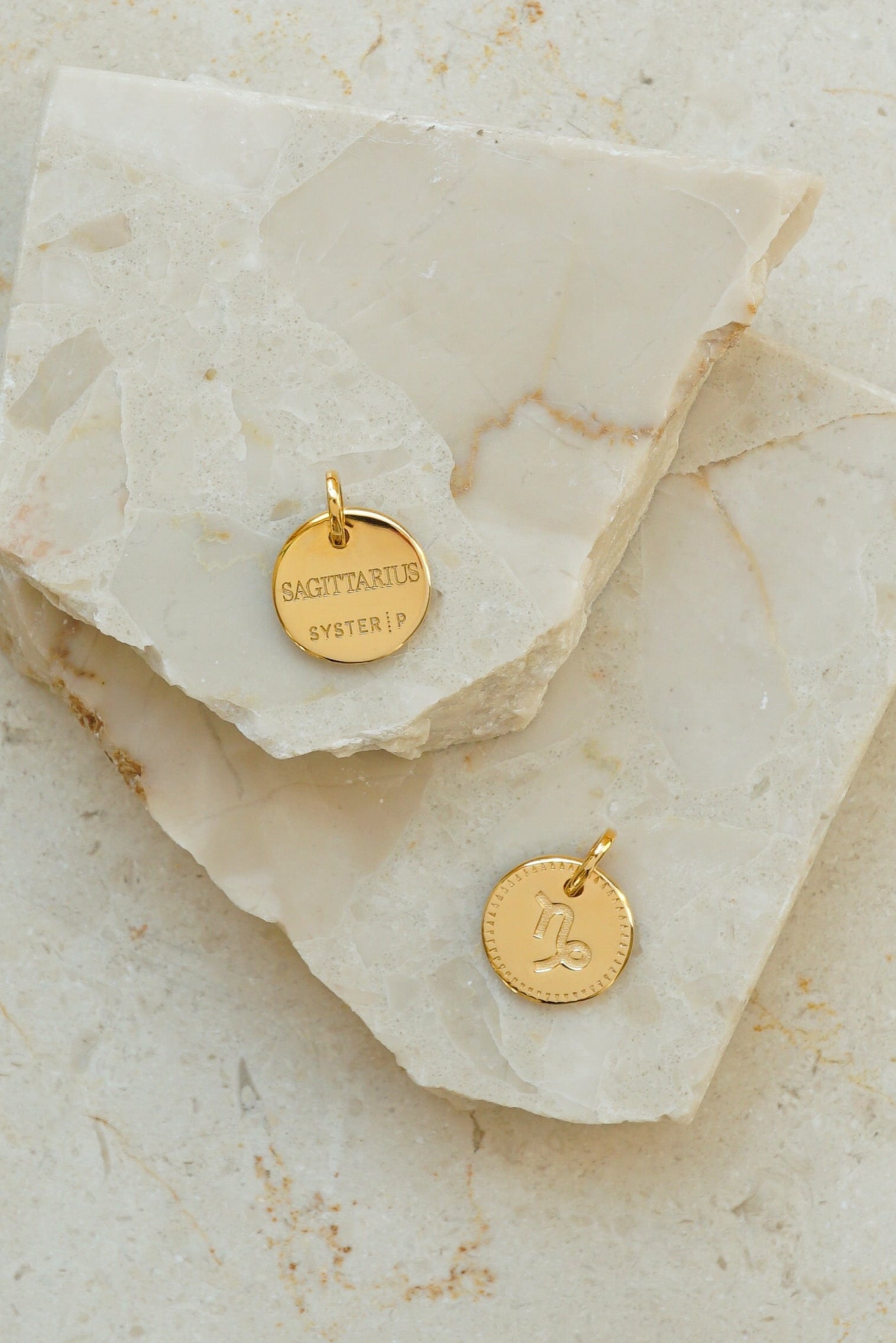 A product shot of two gold Beloved Zodie Pendants on a marble surface, one showing the front with the engraved Capricorn symbol and the other showing the back with the engraved word 'Sagittarius'.