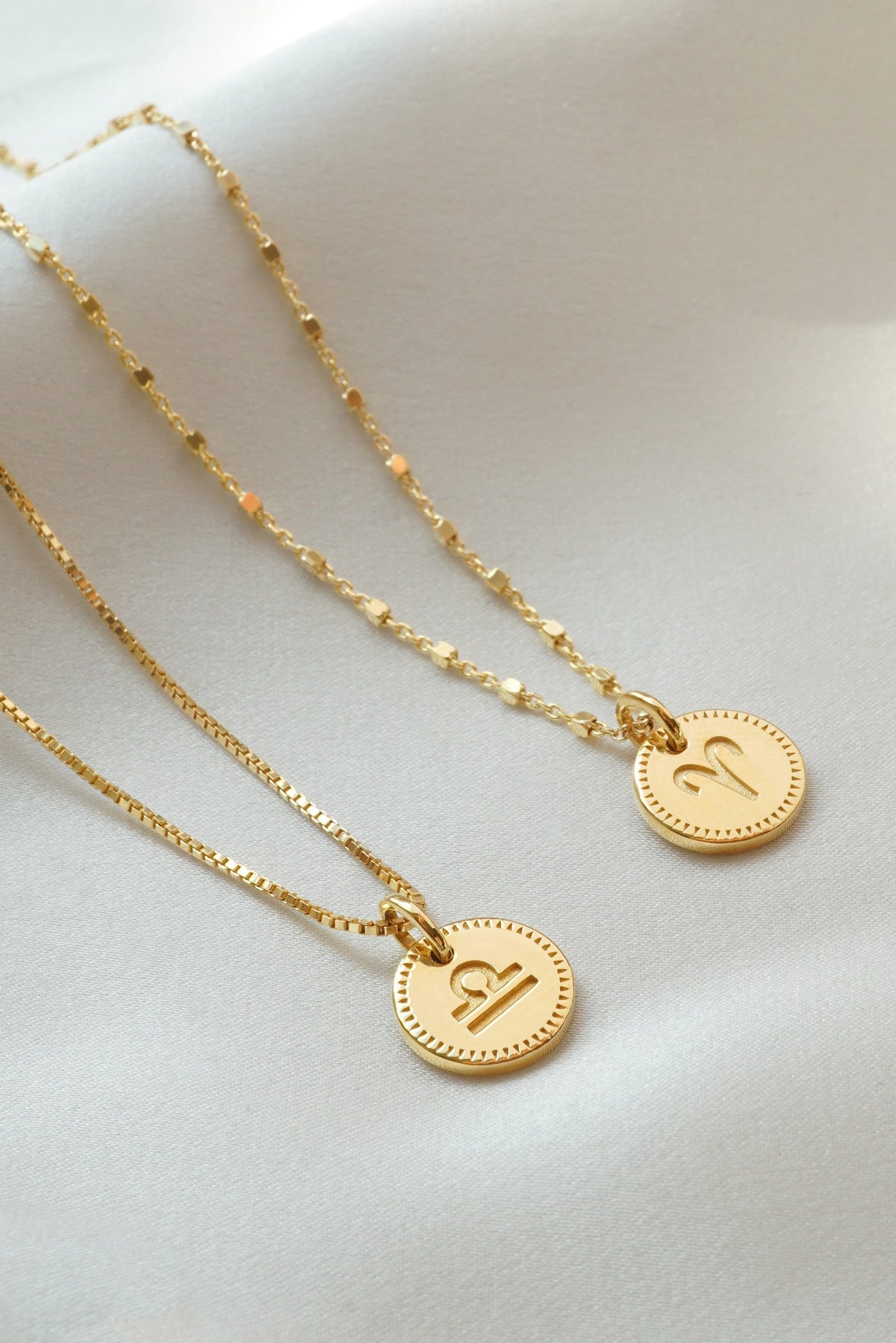 A styling shot of two gold Beloved Zodie Pendants, one engraved with the Libra symbol on a box chain and the other with the Aries symbol on a station chain.