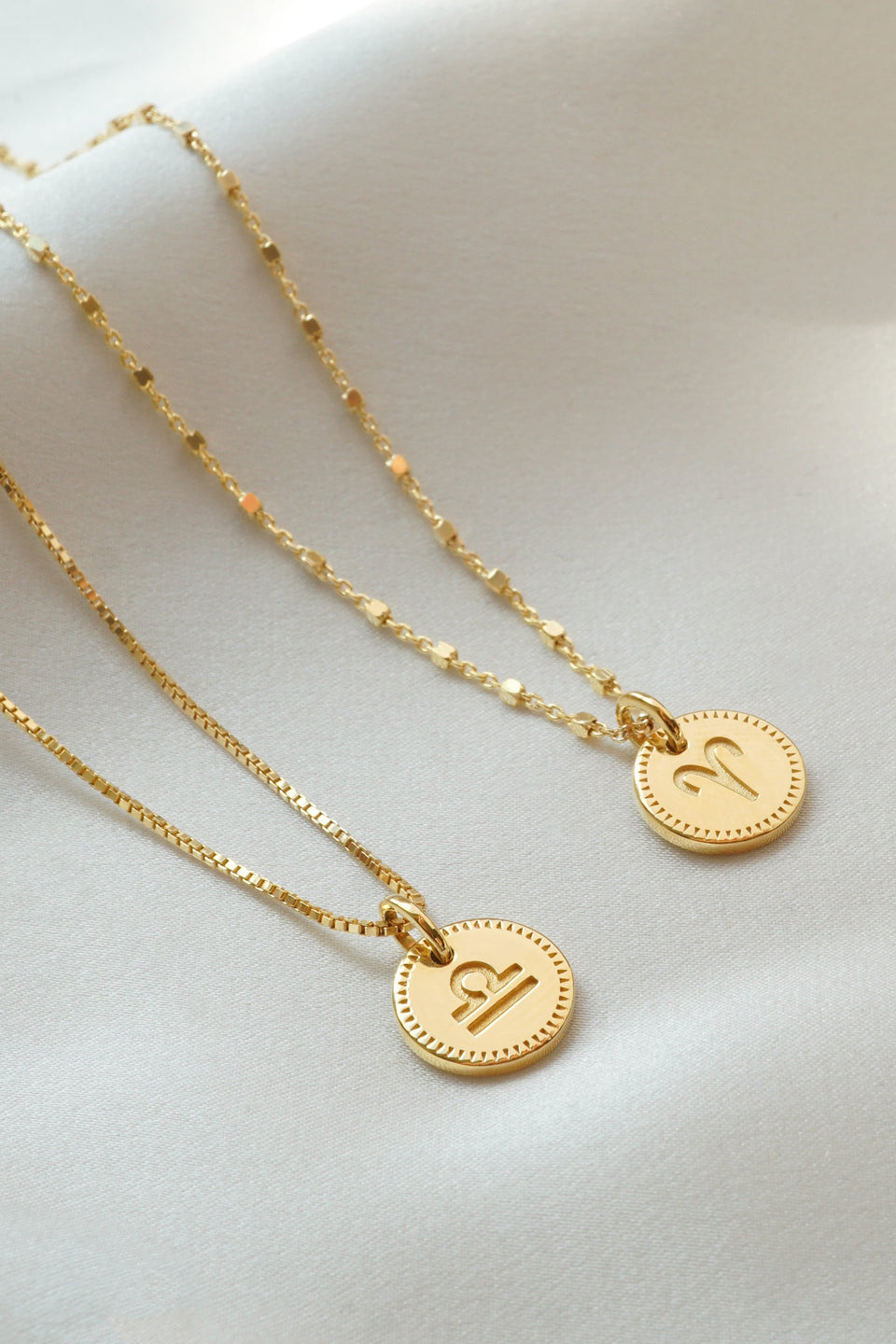 A styling shot of two gold Beloved Zodie Pendants, one engraved with the Libra symbol on a box chain and the other with the Aries symbol on a station chain.