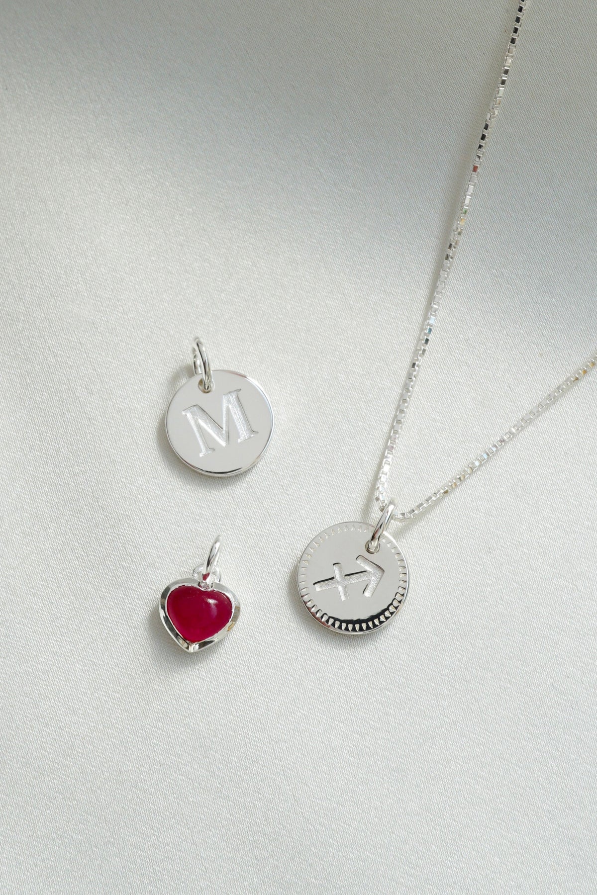 A product shot of a silver box chain necklace with a round Beloved Zodie Pendant featuring the Sagittarius sign, alongside a round silver Beloved Mini Letter 'M' pendant and a small silver Beloved Heart Pendant with a red stone.