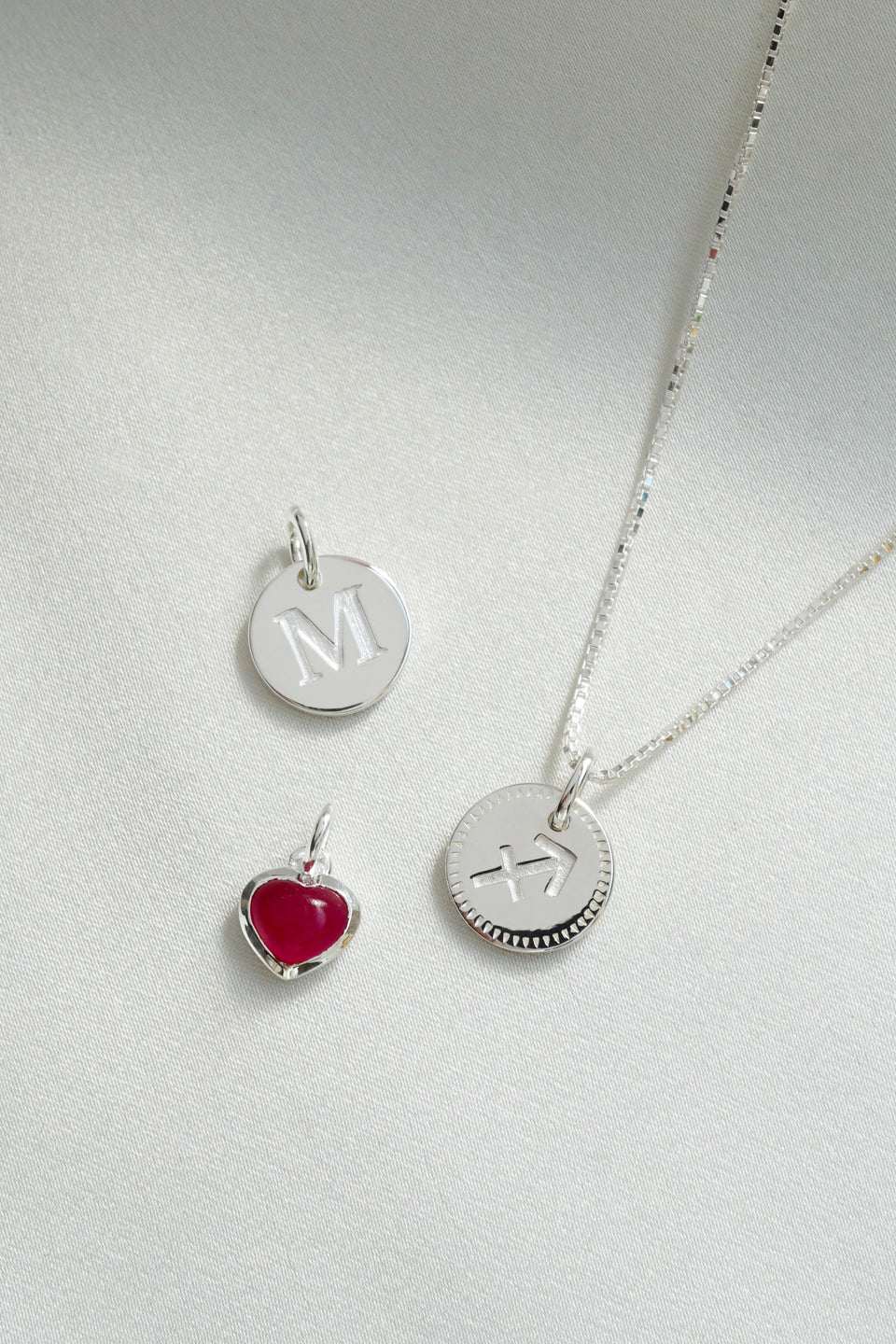 A product shot of a silver box chain necklace with a round Beloved Zodie Pendant featuring the Sagittarius sign, alongside a round silver Beloved Mini Letter 'M' pendant and a small silver Beloved Heart Pendant with a red stone.