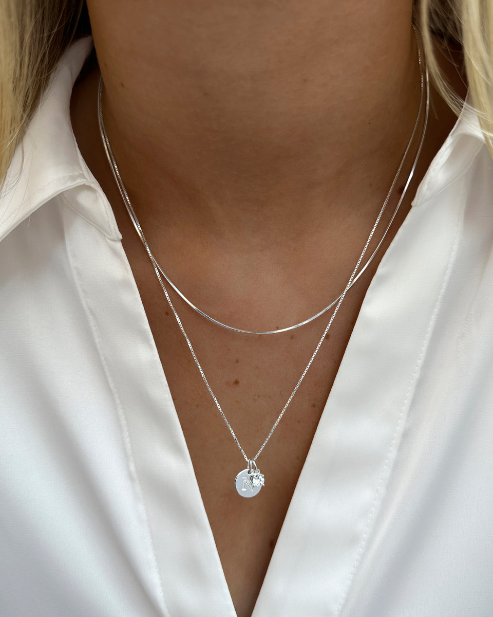 A close-up of a model wearing the Beloved Solo Nova Pendant in silver, styled on a delicate chain with a round initial pendant. The necklace is layered with a flat silver snake chain.