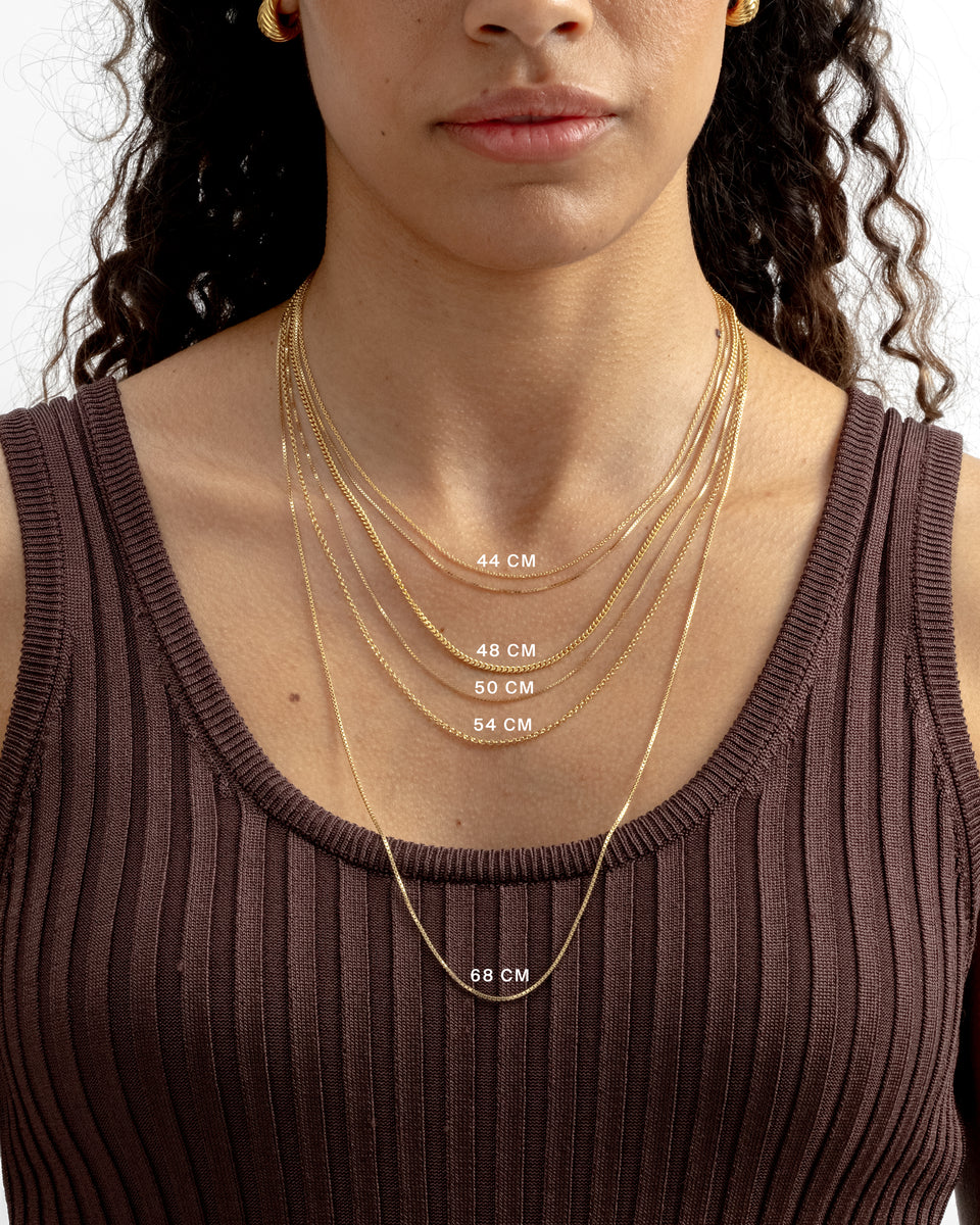 A model showcasing five layered gold necklaces of different styles to illustrate chain lengths, which are marked on the image from 44 cm to 68 cm. The model is also wearing chunky, twisted gold earrings.
