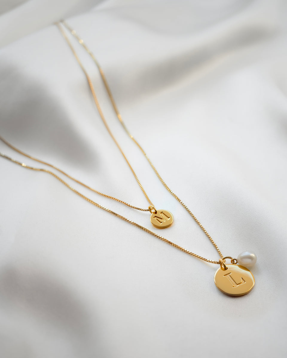 A product shot of the Beloved Long Box Chain in gold, styled with a large 'L' initial pendant and a pearl pendant, layered with a second gold necklace featuring a mini 'M' initial pendant.