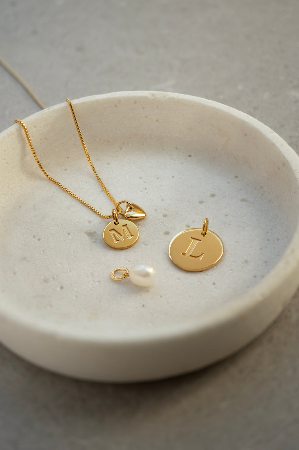A product shot of various gold pendants from the Syster P Beloved collection, including a freshwater pearl pendant, a heart pendant, a small letter 'M' pendant on a box chain, and a larger letter 'L' pendant.