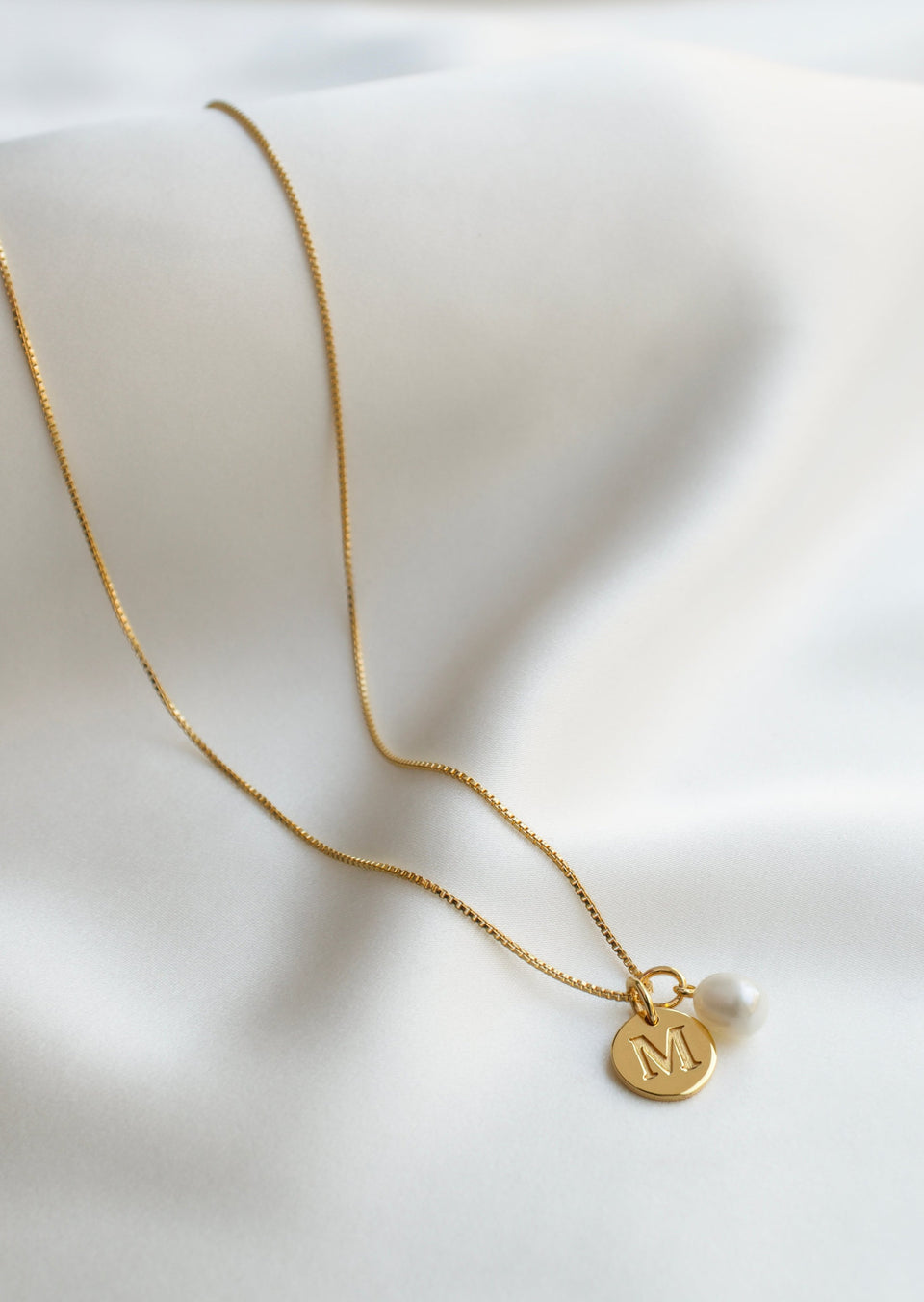 A product shot of a Syster P gold necklace, featuring a medium box chain adorned with a round gold 'M' letter pendant and a single freshwater pearl pendant.
