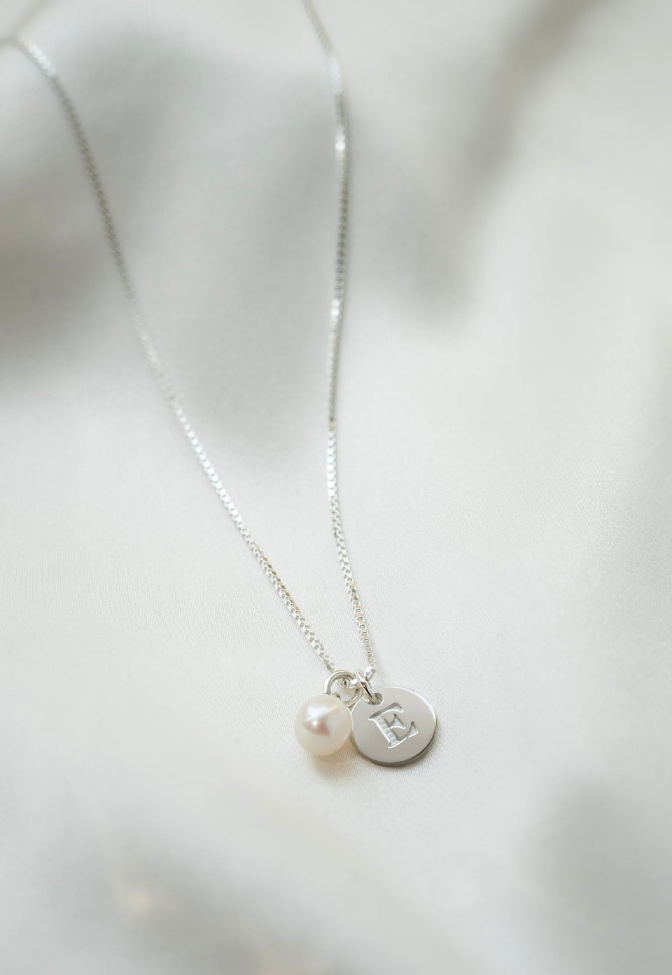 A product shot of a silver necklace featuring a box chain, a white freshwater pearl pendant, and a small, round silver pendant engraved with the letter 'E'.