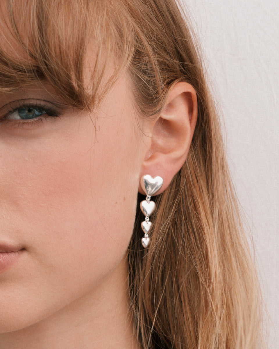 A close-up shot of a model wearing the Darling Earrings in silver, featuring a long pendant made of four cascading, bold hearts.