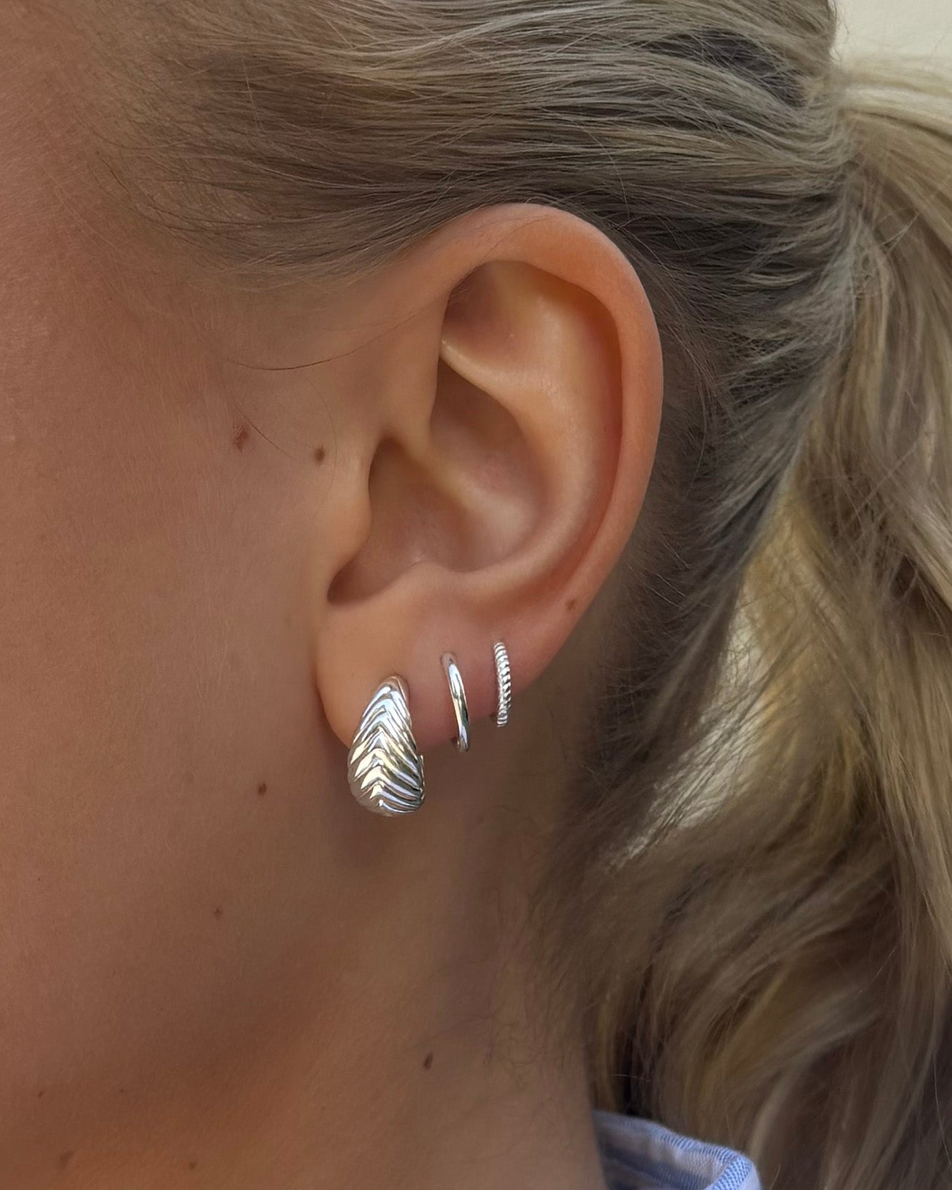 A close-up of a model's ear styled with three silver earrings: the Bolded Little Sis Chevron Earrings, the smooth Beloved Small Fat Hoops, and the textured Beloved Twisted Small Hoops.