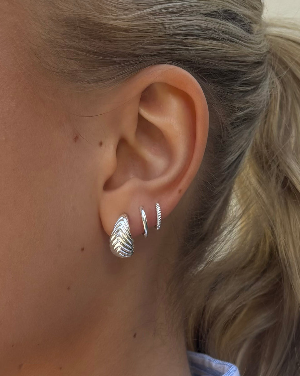 A close-up of a model's ear styled with three silver earrings: the Bolded Little Sis Chevron Earrings, the smooth Beloved Small Fat Hoops, and the textured Beloved Twisted Small Hoops.