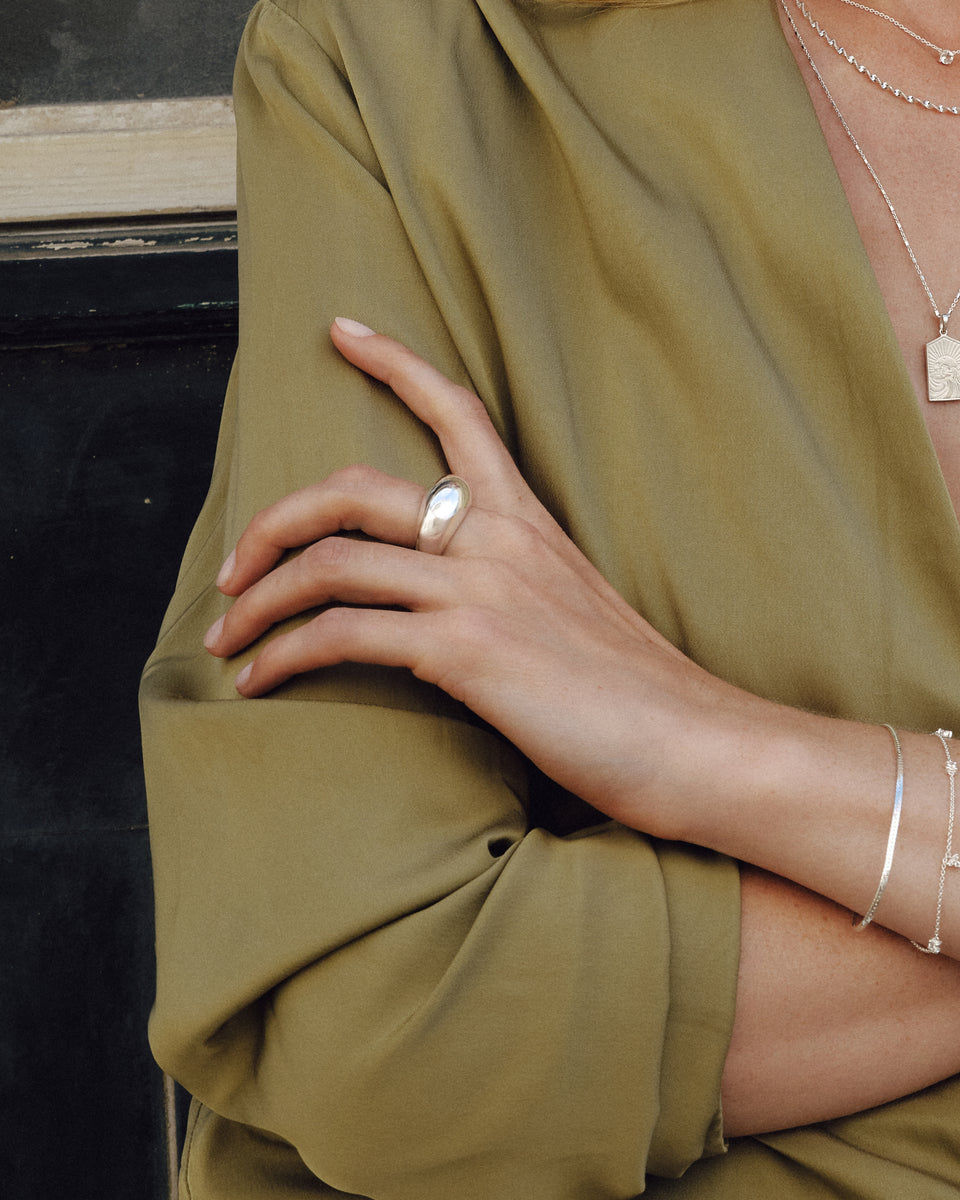 A close-up shot of a model wearing the Bolded Big Ring in silver, a statement dome-shaped ring. She is also wearing several layered silver necklaces and two silver bracelets: the flat Herringbone Bracelet and the delicate Celestia Bracelet.