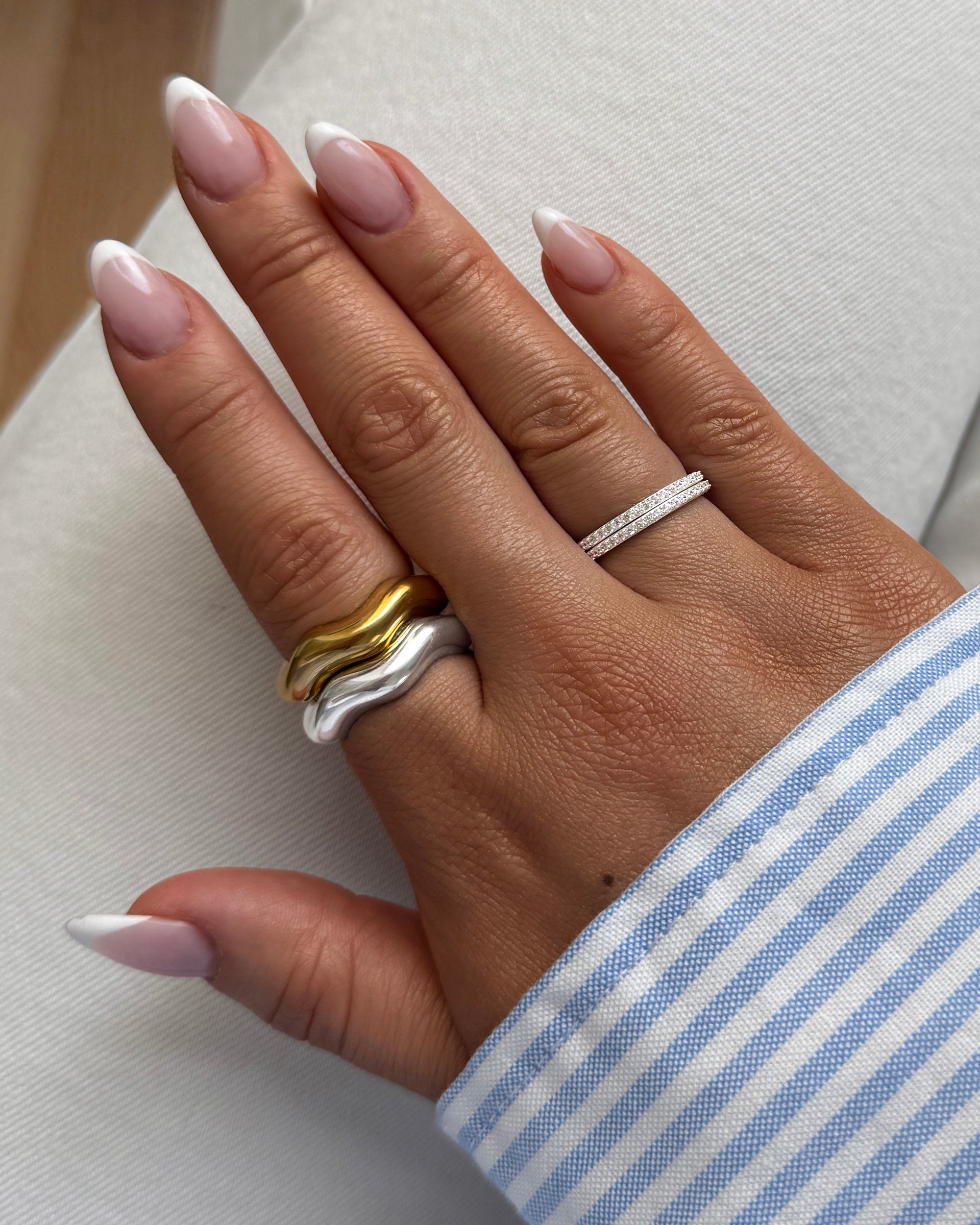 A close-up of a model's hand wearing the Bolded Wavy Ring in silver and gold stacked together, alongside a thin silver ring with sparkling stones.