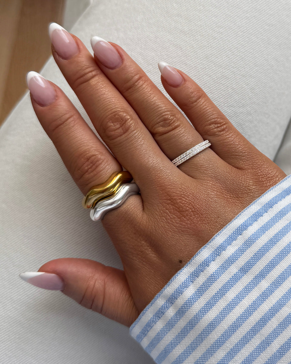 A close-up of a model's hand wearing the Bolded Wavy Ring in silver and gold stacked together, alongside a thin silver ring with sparkling stones.