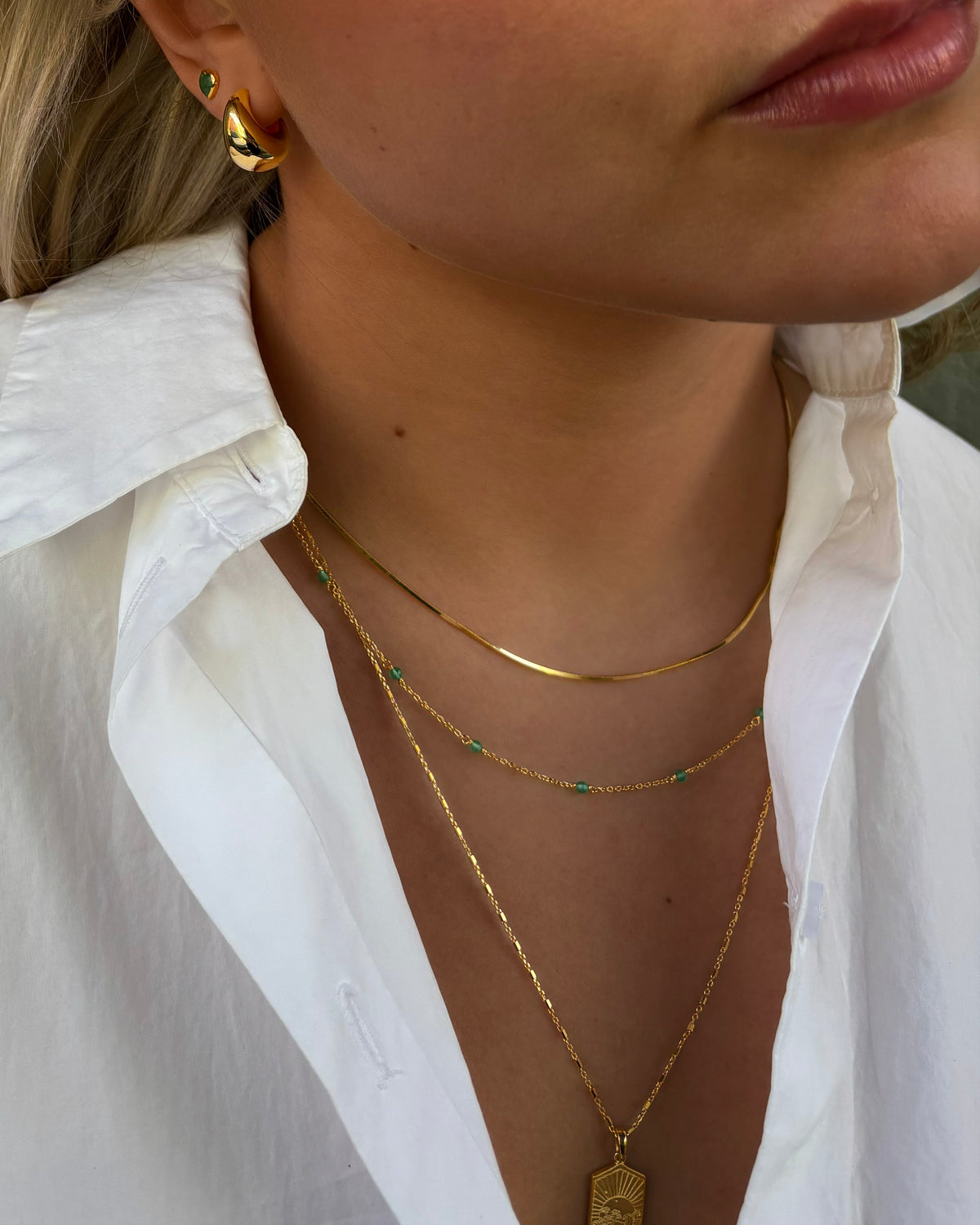 Close-up of a model wearing the Santa Monica Necklace Gold, a delicate gold chain with green onyx beads, layered with a gold chain and a gold pendant necklace, complemented by bold gold earrings and a green heart-shaped stud.