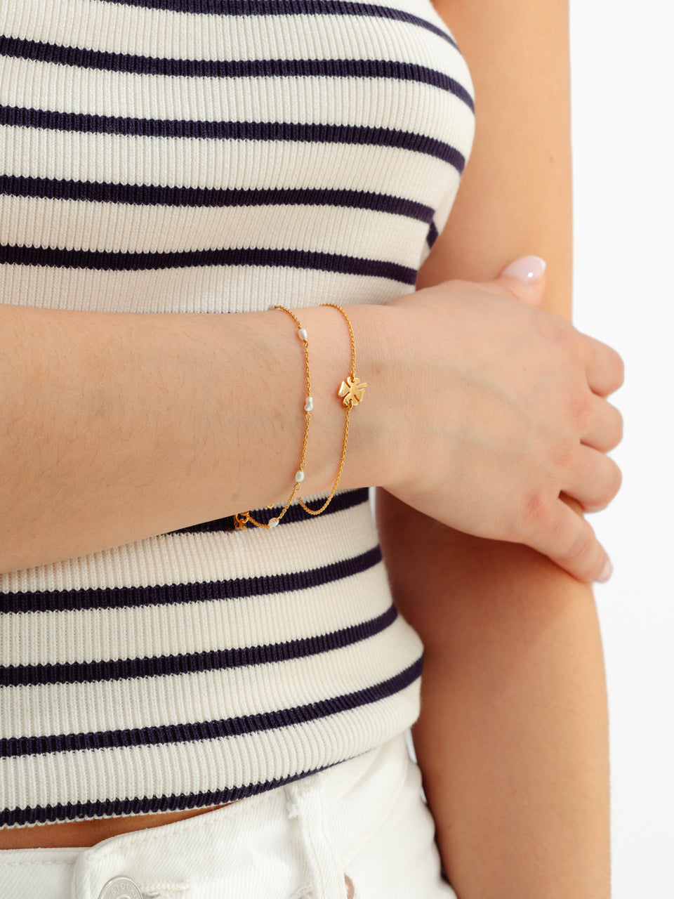 A model wears the Bring Me Luck Bracelet in gold, featuring a thin chain and a four-leaf clover charm, layered with another fine gold chain bracelet with small pearls.