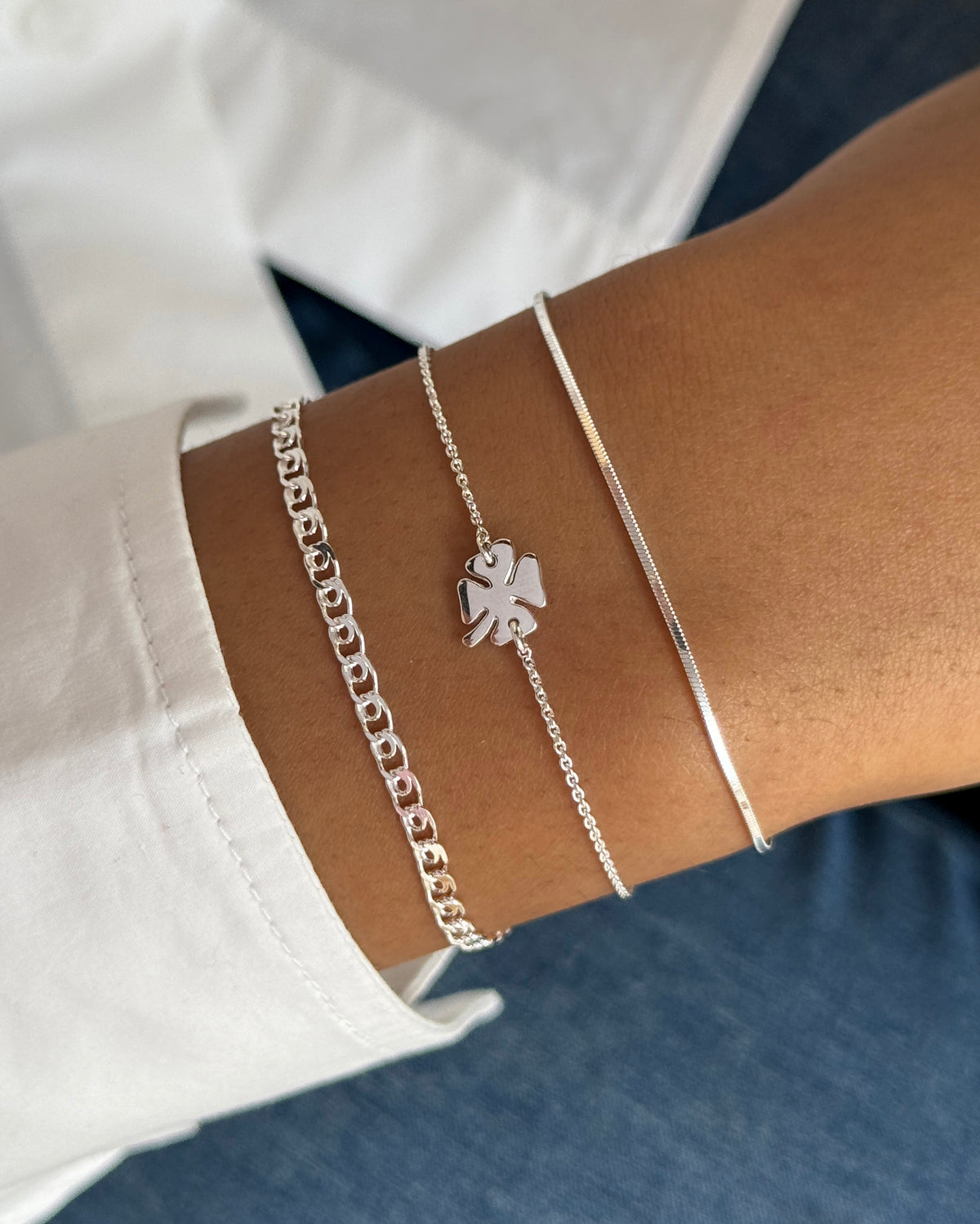 A close-up shot of a model's wrist styled with three stacked Syster P bracelets in silver: the flat Sheer Mirror snake chain, the delicate Bring Me Luck chain with a four-leaf clover charm, and the classic Sheer Love curb chain.