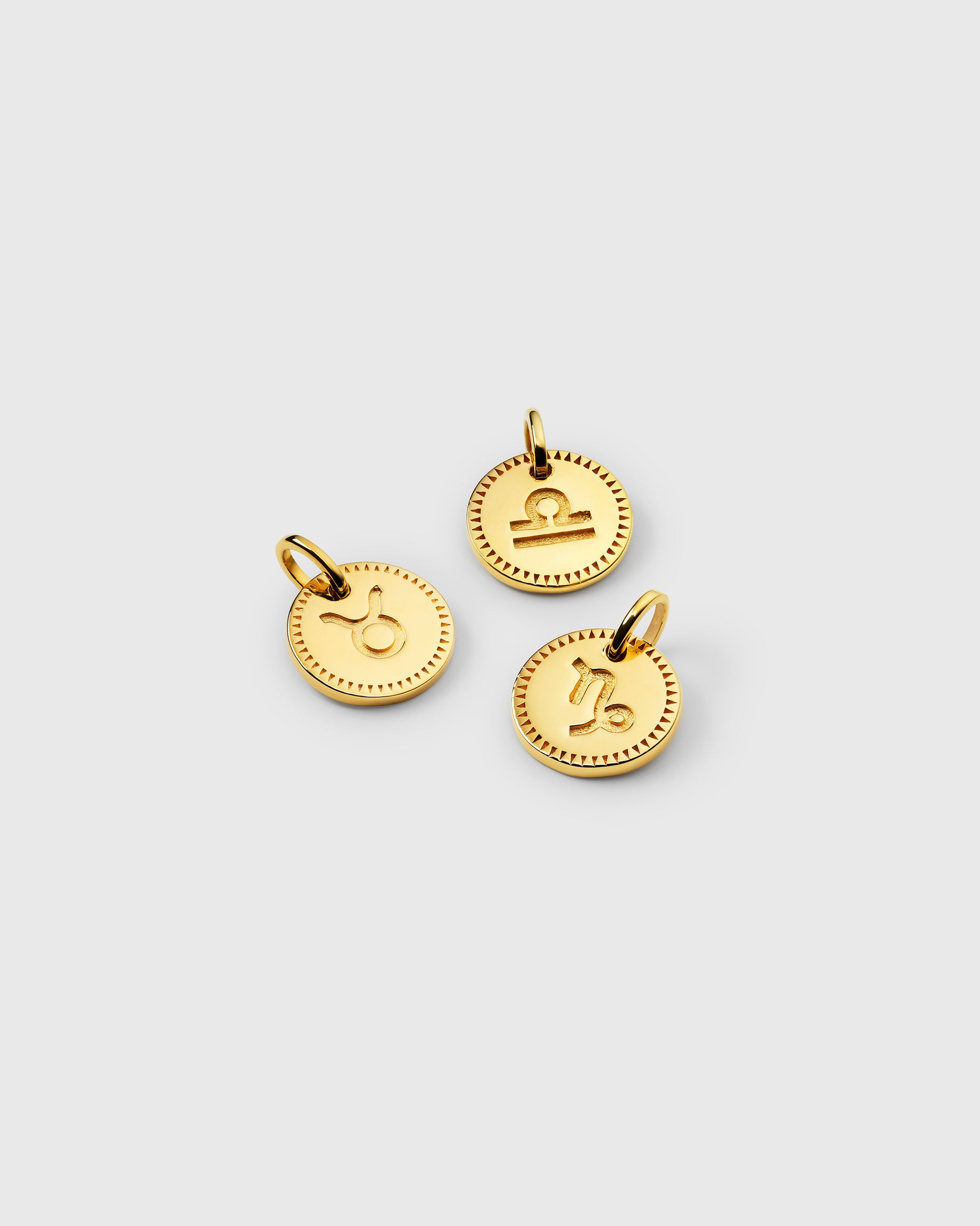A product shot of three Beloved Zodie Pendants in gold, displaying the Taurus, Libra, and Capricorn signs. Each circular charm features an engraved zodiac symbol in the center, framed by a decorative border.