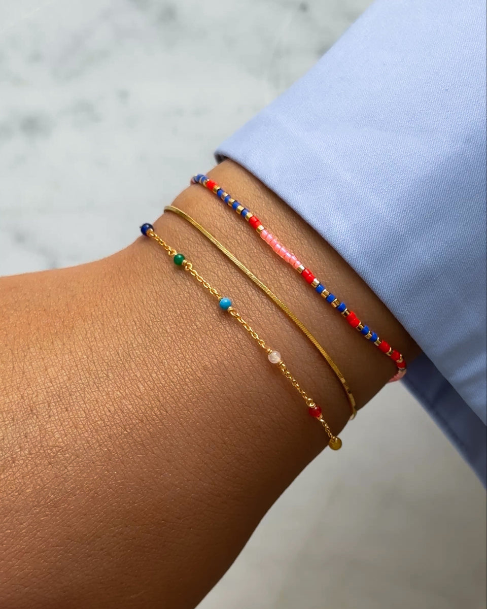 A model wears a stack of three gold bracelets, including the Code Bracelet Gold Just Breathe with red, blue, and pink morse code beads, a flat gold snake chain, and a delicate chain with small multi-colored beads.