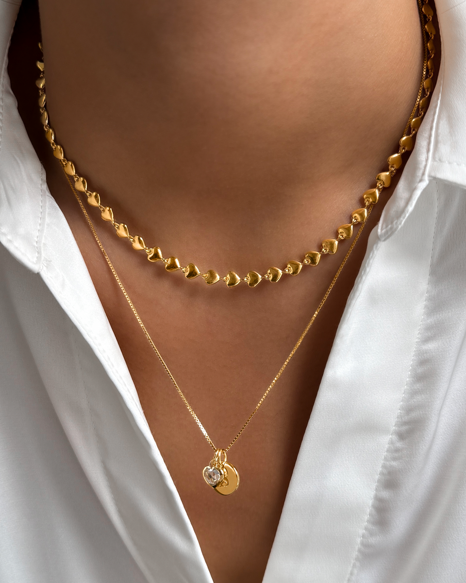 A close-up shot of a model wearing the gold Darling Necklace, a chain of small interconnected hearts, layered with a finer gold box chain necklace featuring a crystal heart pendant and a small letter charm.