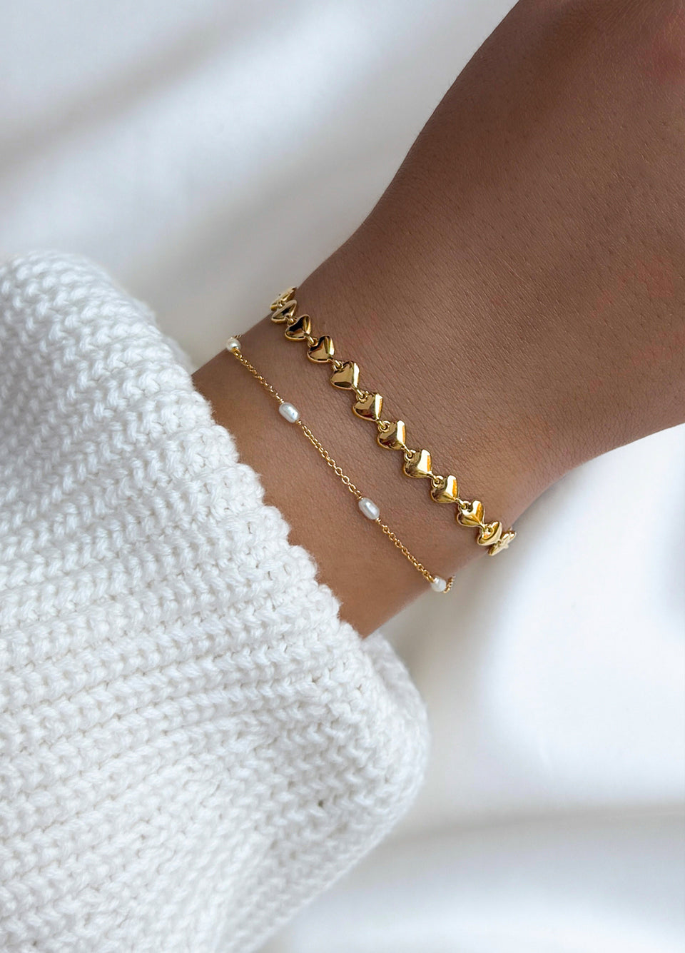 A close-up of a model wearing the Treasure Multi Pearl Bracelet in gold, featuring delicate freshwater pearls, styled with a second gold bracelet made of linked hearts.