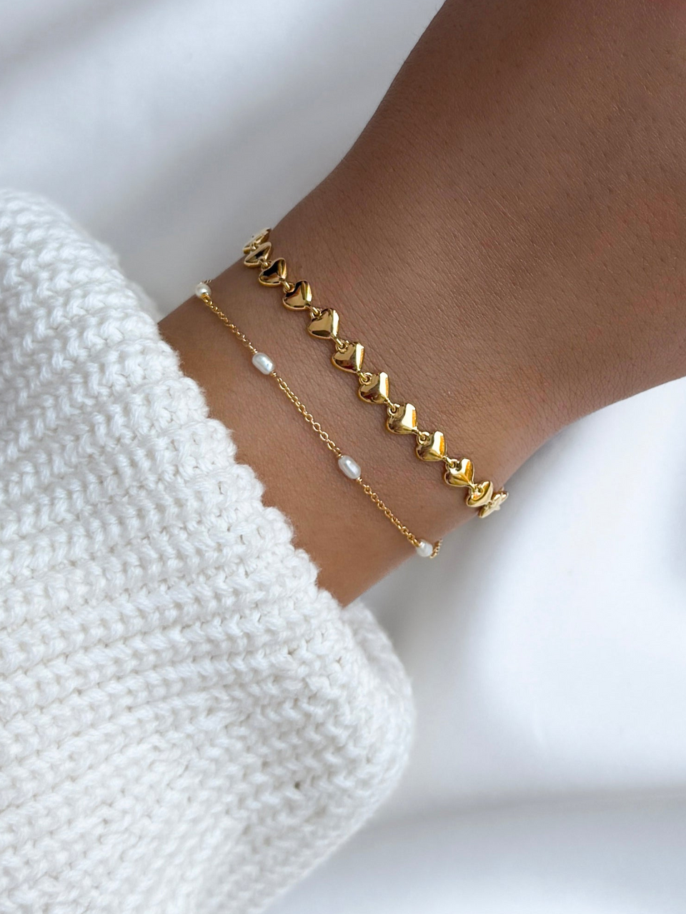 A close-up of a model's wrist, styled with two layered gold bracelets: the Darling Bracelet featuring a chain of hearts, and the delicate Treasure Multi Pearl Bracelet.