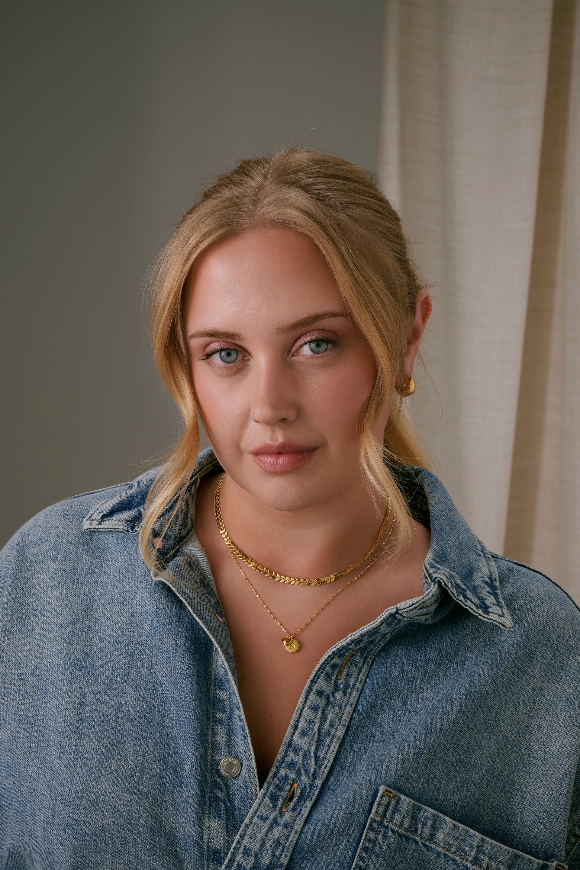 A model wearing layered gold necklaces, including the Sheer Station Necklace with a Beloved Mini Letter 'S' pendant and a gold Beloved Heart pendant.