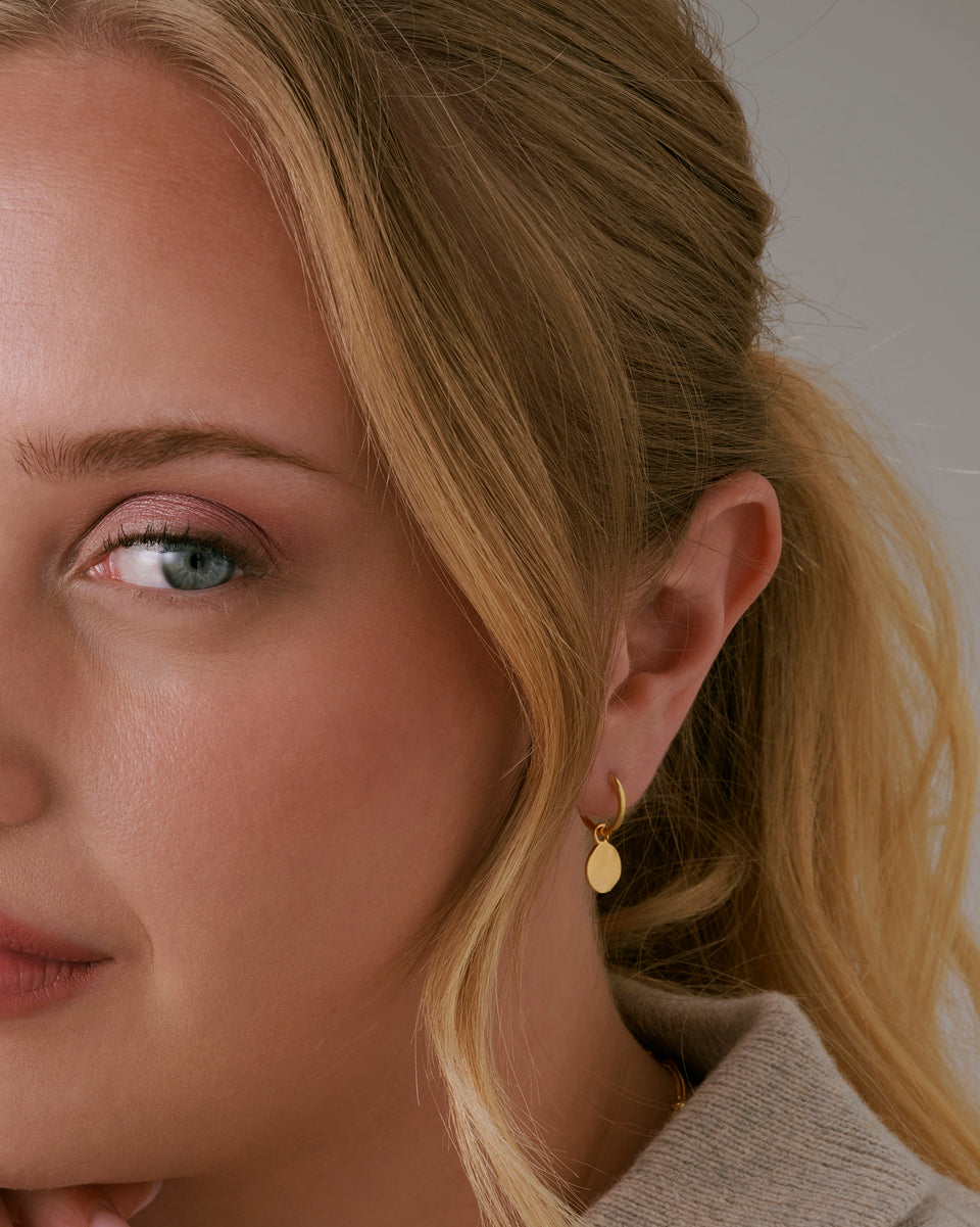 A close-up shot of a model wearing the Minimalistica Hammered Earrings in gold, featuring a small hoop with a dangling, round pendant with a hammered surface.