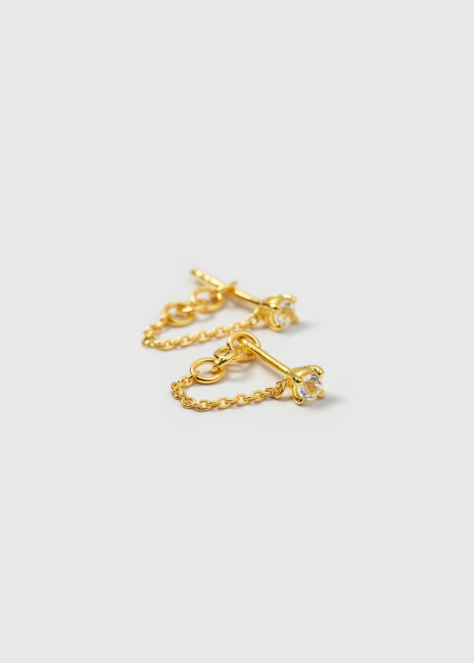 A product shot of the Minimalistica Solo Nova Earrings in gold, featuring small studs with white topaz stones and a delicate chain that connects the stud to the earring back.