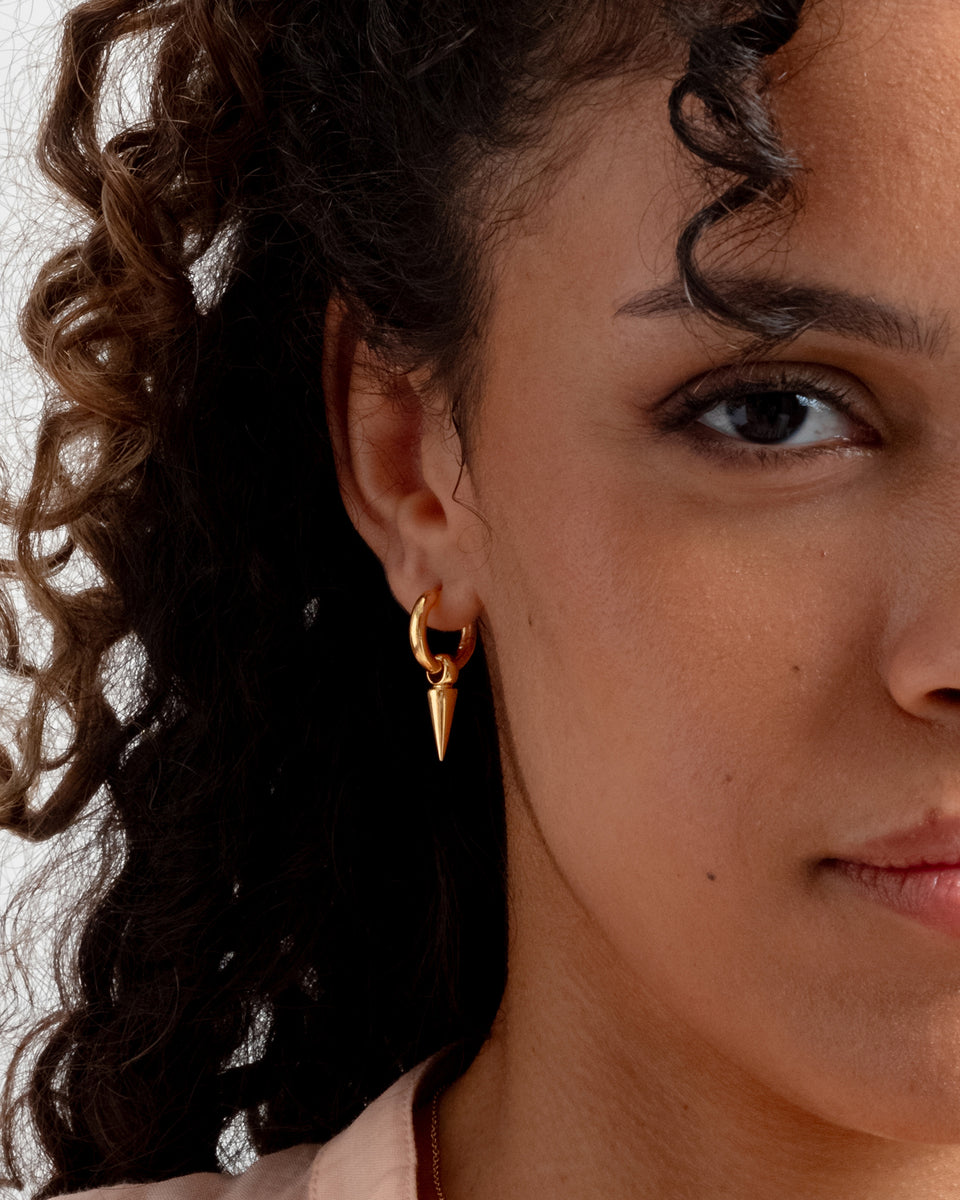 A close-up shot of a model wearing the Juno Hoops in gold, a hoop earring with a dangling spike charm.