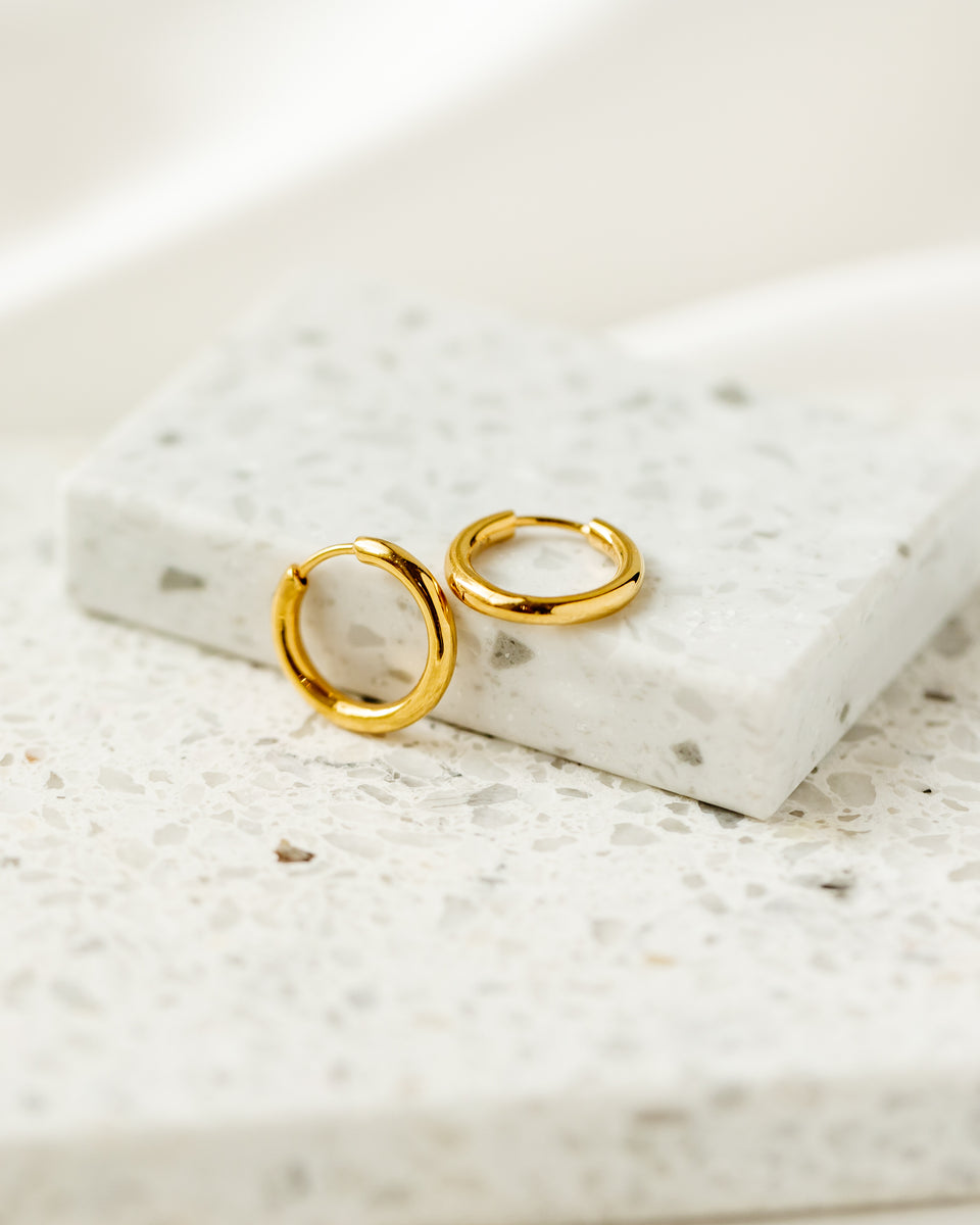 A product shot of the Beloved Fat Medium Hoops in gold, resting on a white terrazzo block that highlights their classic, chunky design.