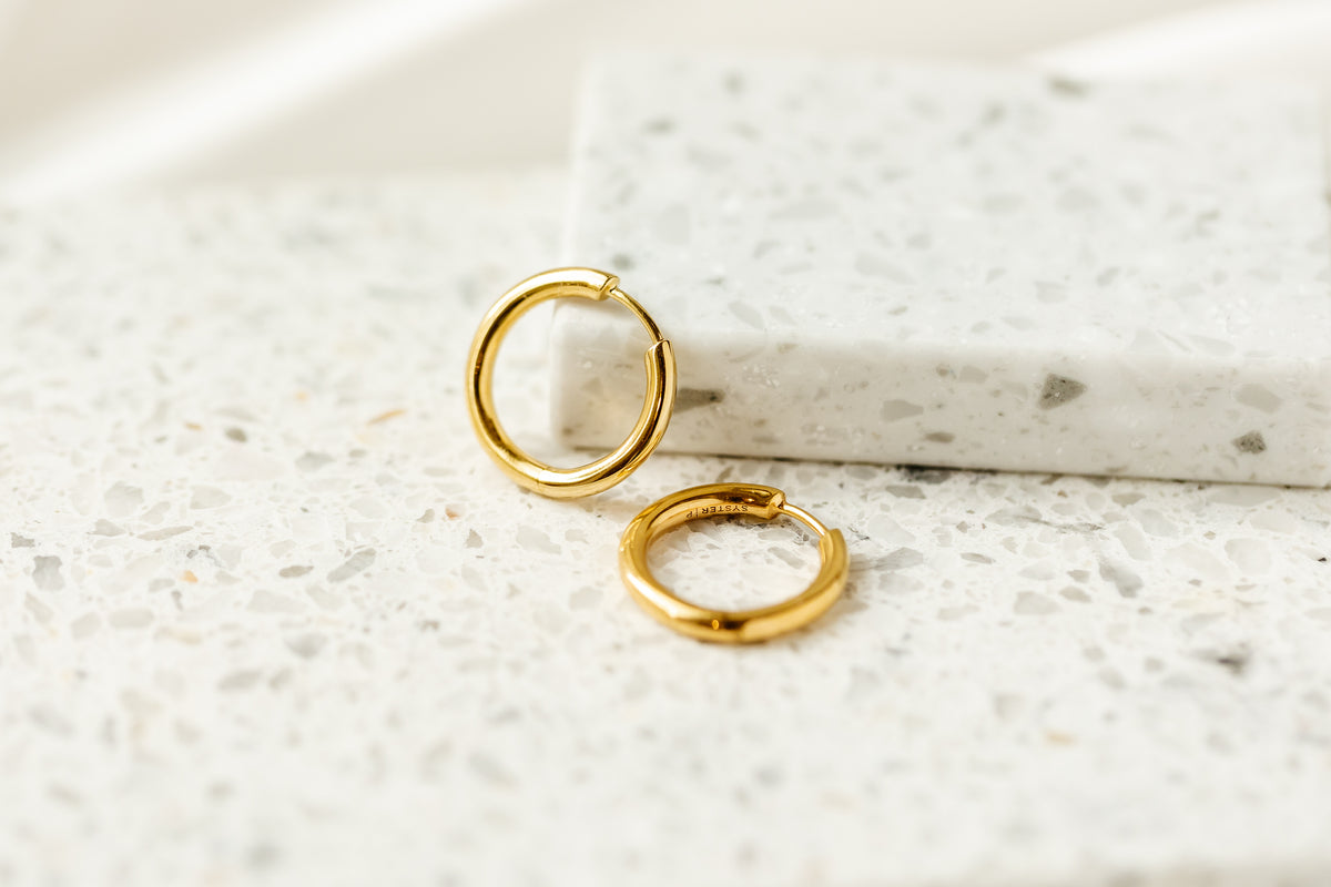 A product shot of the Beloved Fat Big Hoops in gold, a pair of classic, thick hoop earrings resting on a white terrazzo surface.