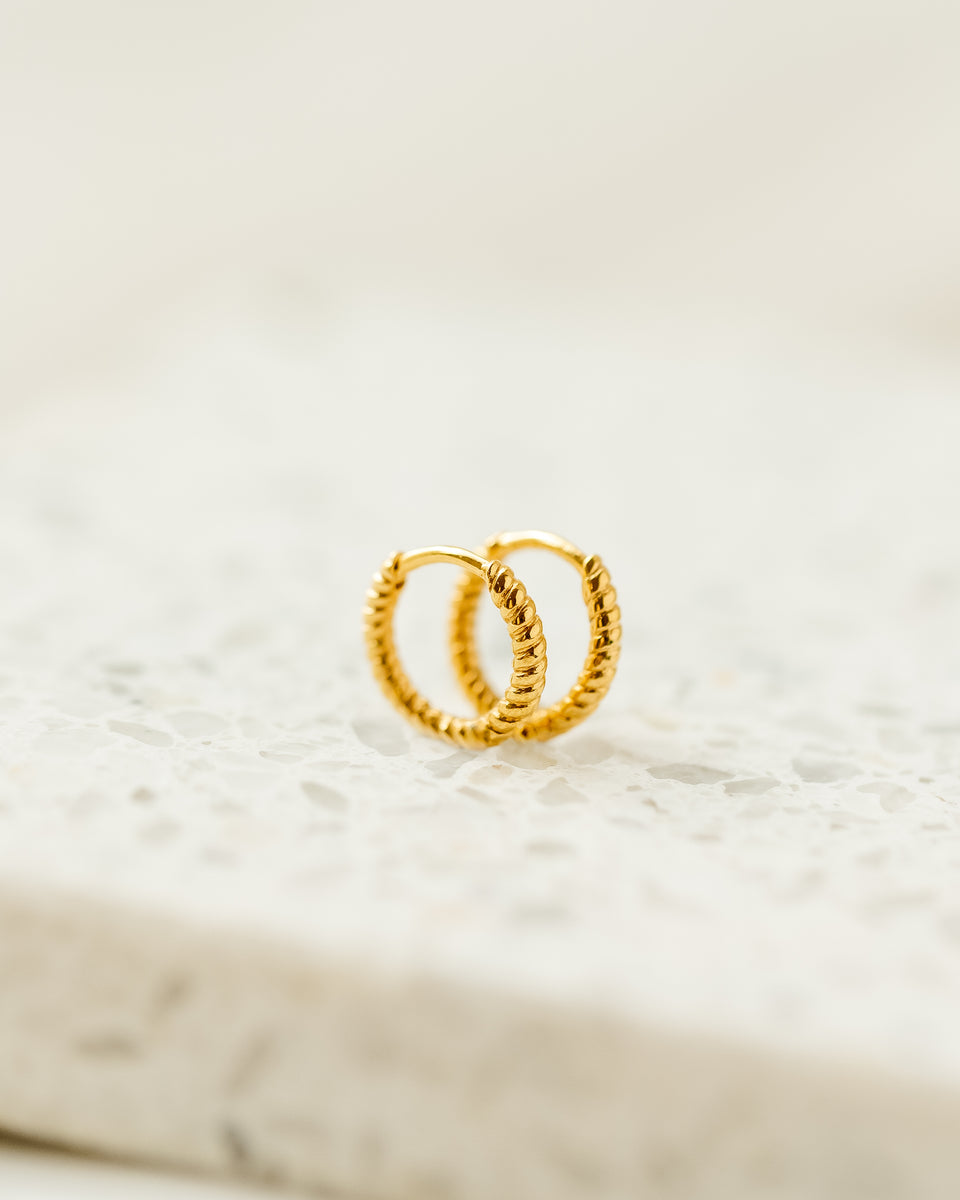 A product shot of the Beloved Twisted Small Hoops in gold, resting on a light surface that highlights their intricate twisted design.