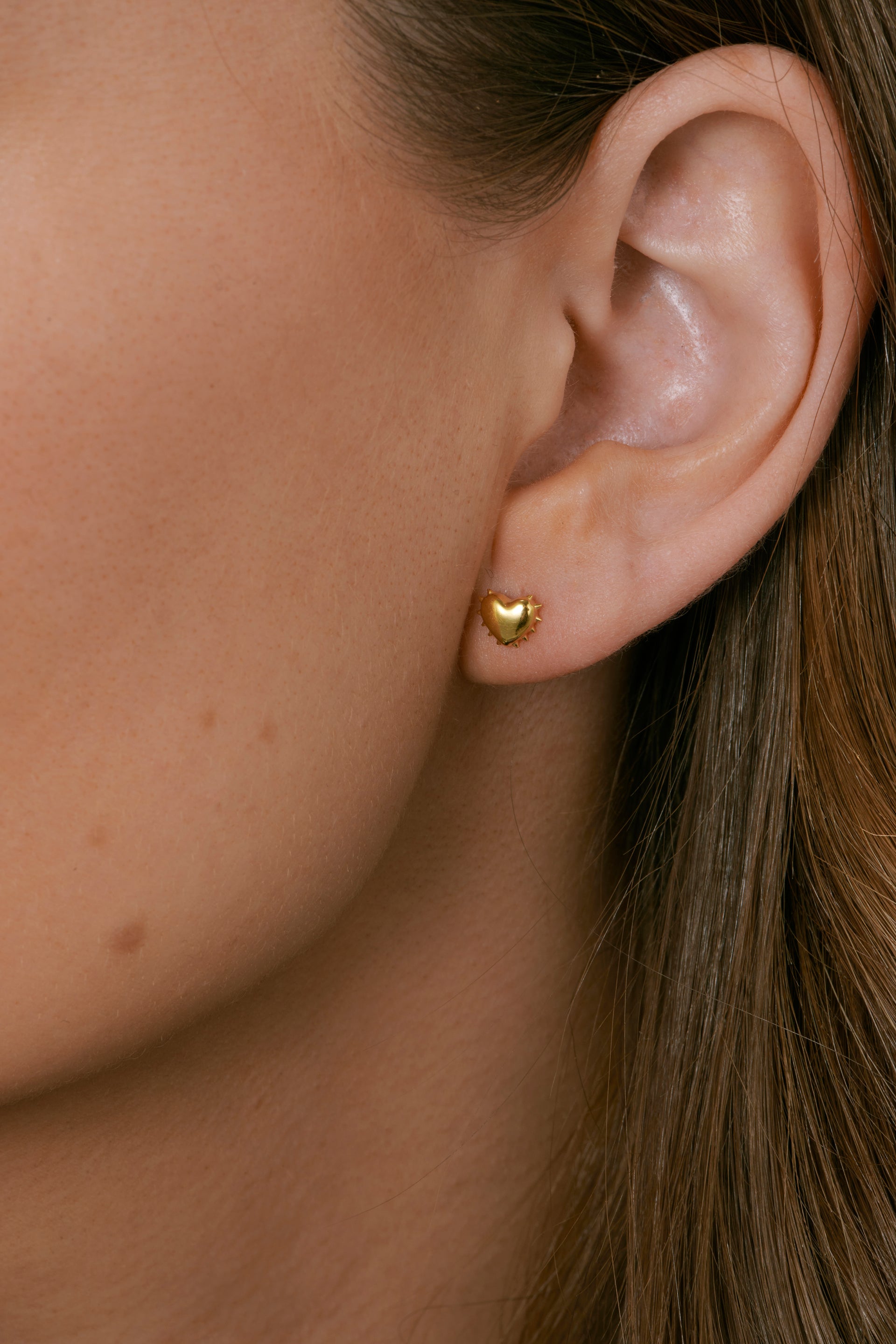 A close-up image of a model wearing the True Love Spike Heart Studs in gold, showing the small heart-shaped earring with spike details on her earlobe.