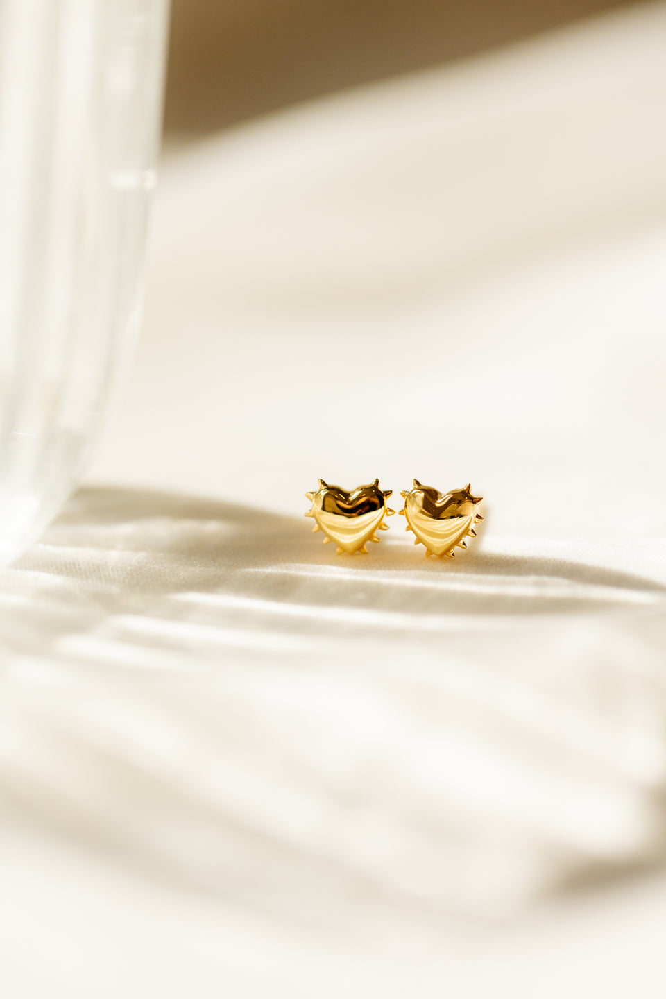 A close-up image of the True Love Spike Heart Studs in gold, detailing their unique heart shape with a spiked trim as they rest on a soft, light-colored fabric.