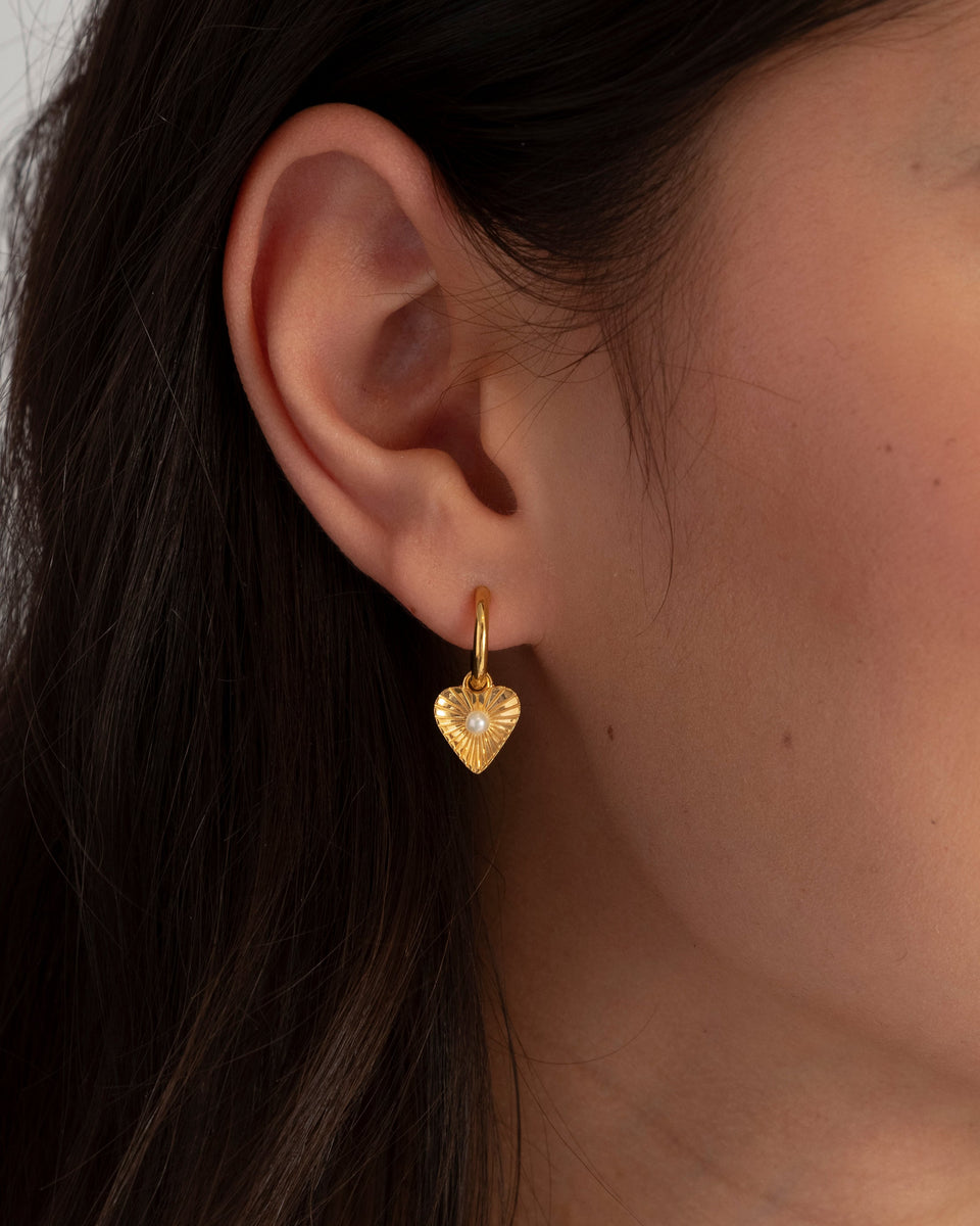 A close-up shot of a model wearing the Aurelia Earrings in gold, featuring a dangling heart-shaped pendant with a textured ray design and a central freshwater pearl.