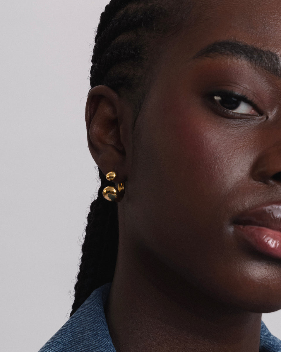 A close-up shot of a model wearing the Bolded Drops Earrings in gold, showcasing their unique design of two rounded, drop-like shapes.