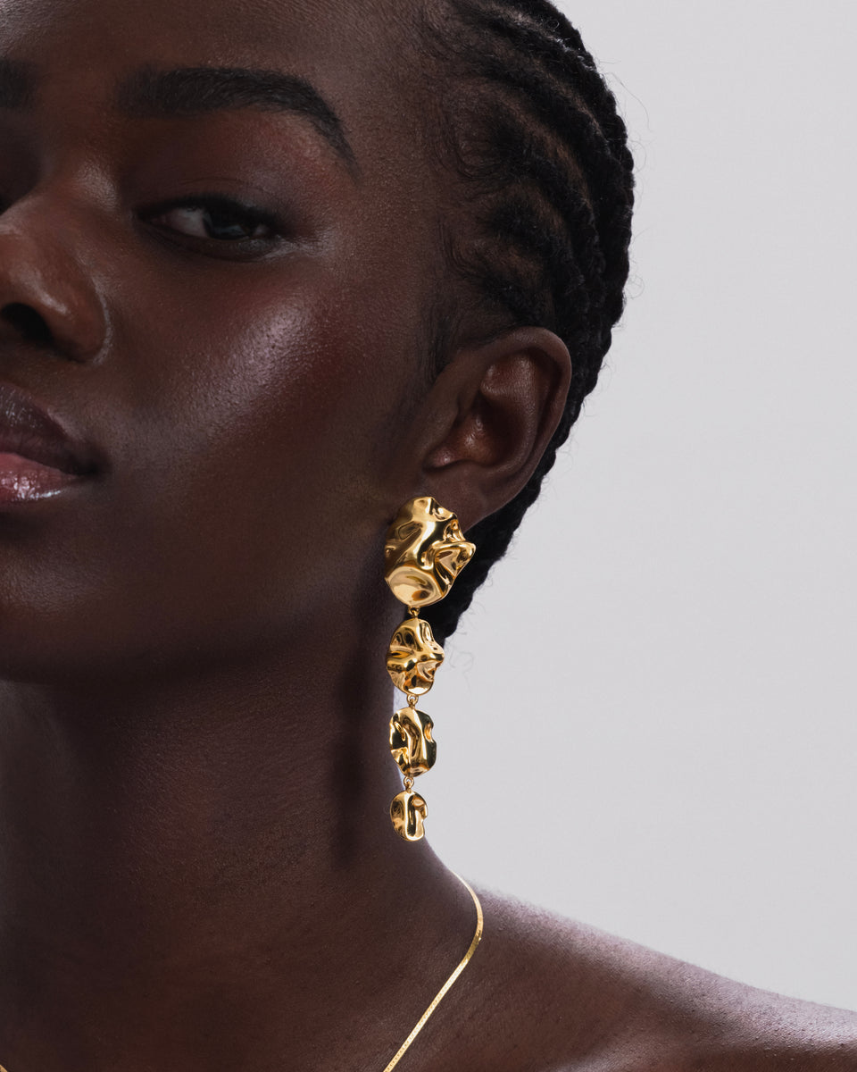 A close-up shot of a model wearing the Moulded Long Single Earring in gold, which features a long, dangling design with an organic, irregular surface. She is also wearing a thin gold snake chain necklace.