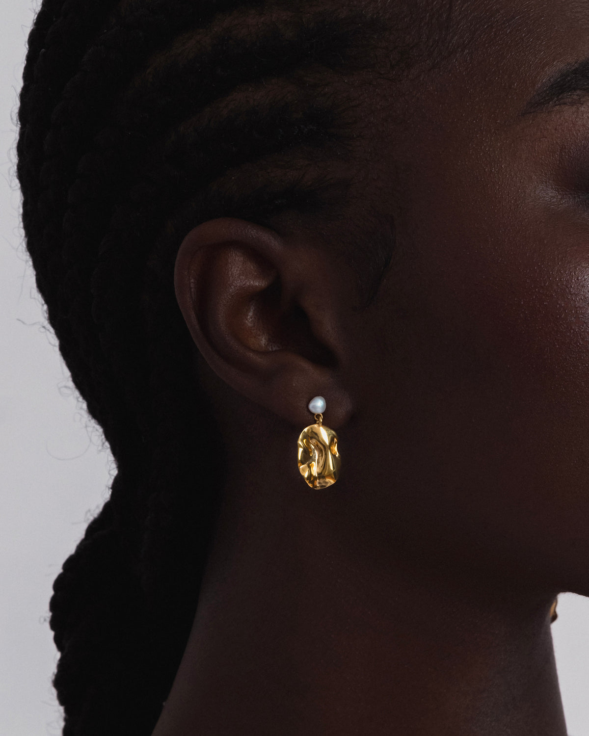 A close-up shot of a model wearing the Moulded Short Earring in gold, featuring a freshwater pearl stud and a dangling, textured pendant.