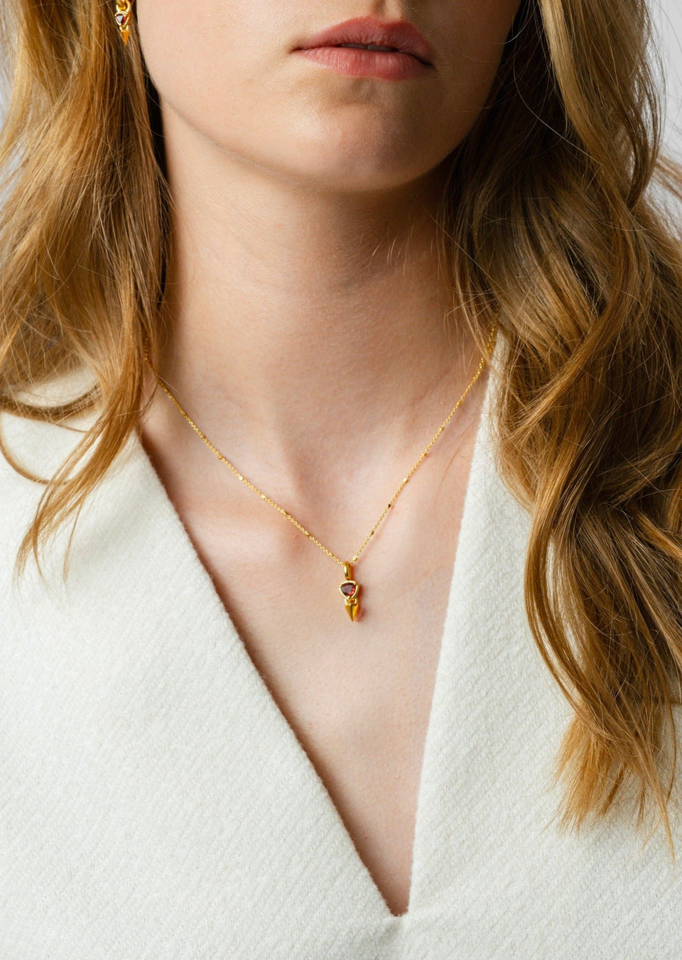A close-up shot of a model wearing the Mrs H Spike Necklace in gold, featuring a pendant with a spike and a red garnet, along with the matching earring.