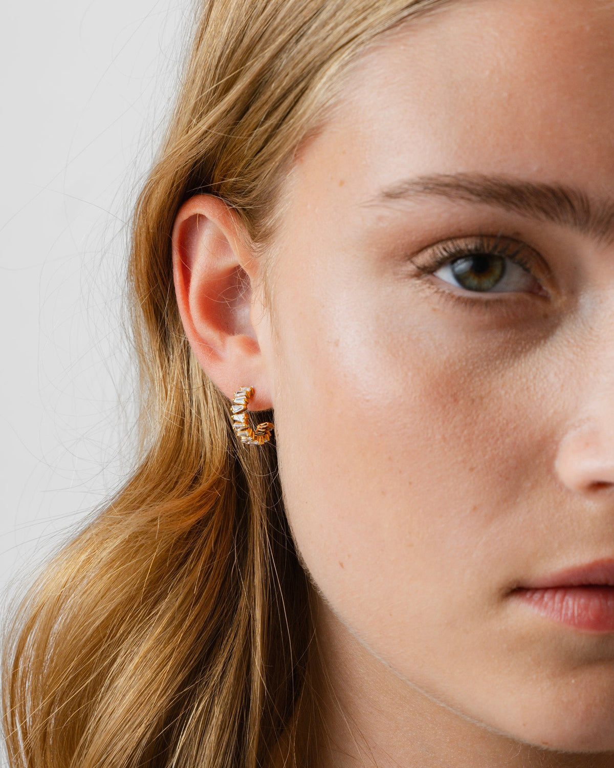 A close-up shot of a model wearing the Iggy Earrings in gold, featuring a hoop design adorned with tapered baguette-cut cubic zirconias.