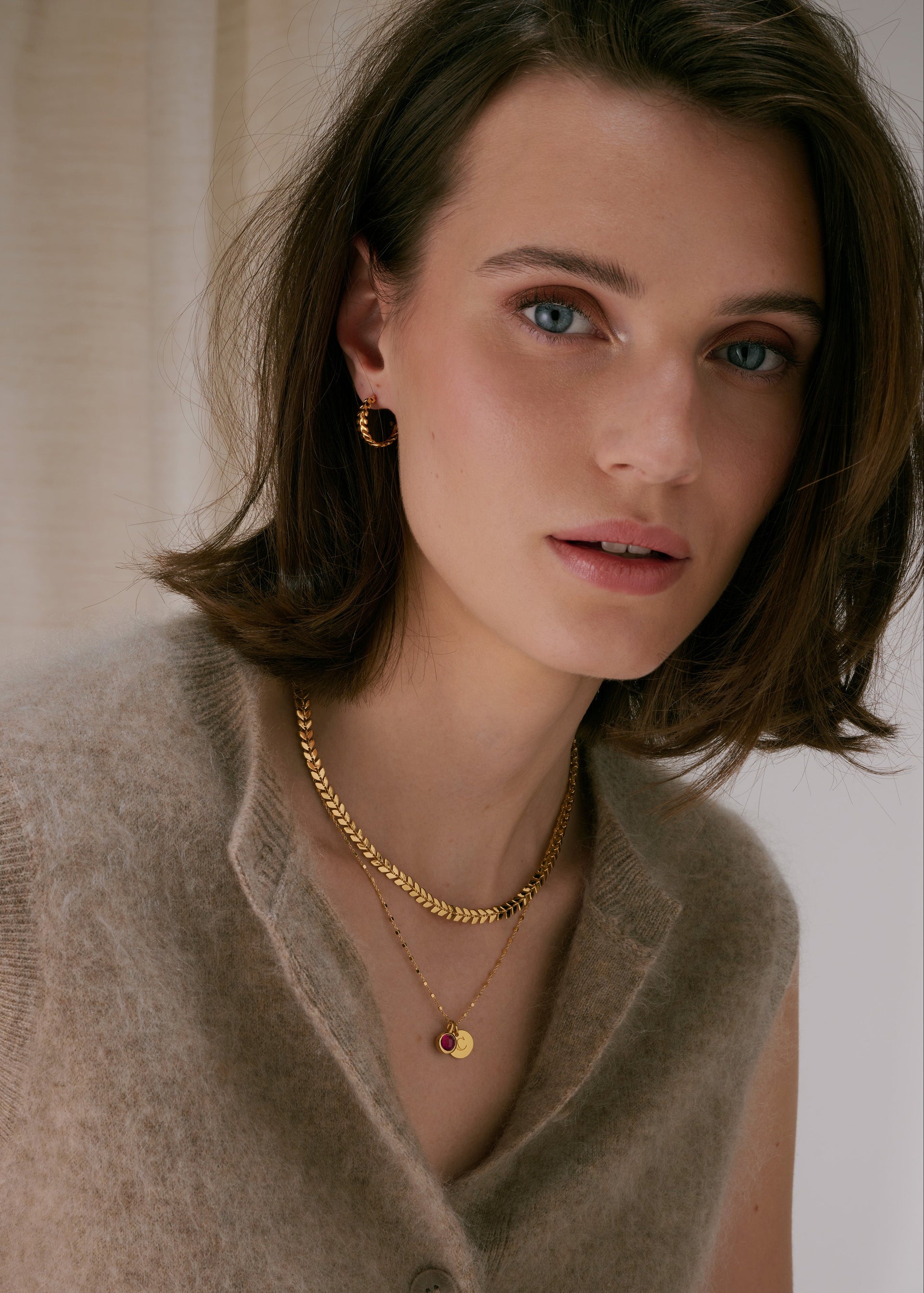 Gold hoop earrings with a leaf-like pattern, paired with two layered gold necklaces—one thick chain with a leaf pattern, and one fine chain with a pendant featuring a ruby, July's birthstone, and an initial charm.