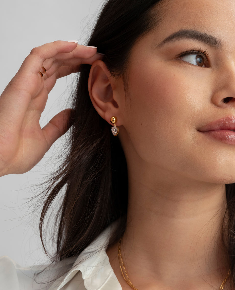 A product shot of the Katie Earrings in gold, featuring small hoops with a dangling, drop-shaped cubic zirconia stone.