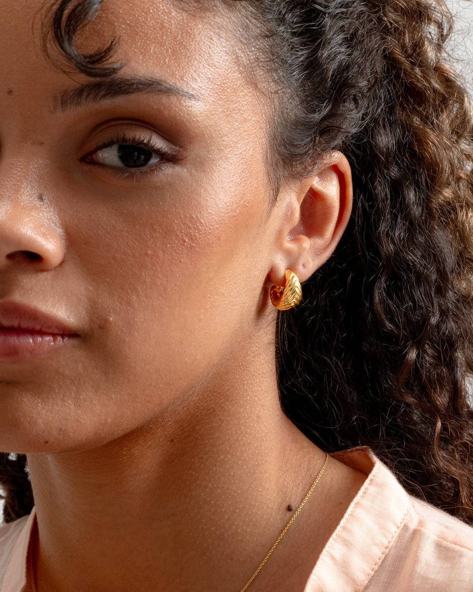 A close-up on a model wearing the Bolded Little Sis Chevron Earrings in gold, showing off their chunky design and chevron pattern. A delicate gold chain necklace is also worn.