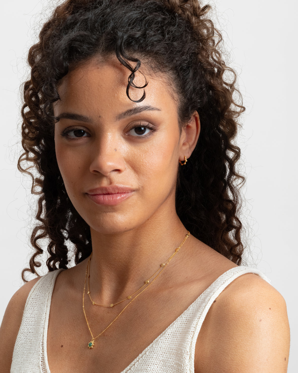 A model styles the Florentina Small Hoops in gold, featuring a unique wavy pattern, with a layered set of fine gold chain necklaces.