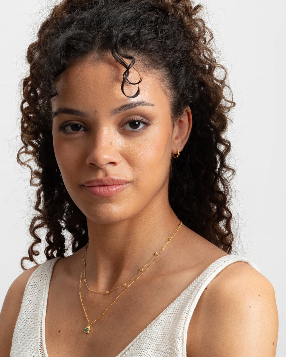 A model styles the Florentina Small Hoops in gold, featuring a unique wavy pattern, with a layered set of fine gold chain necklaces.