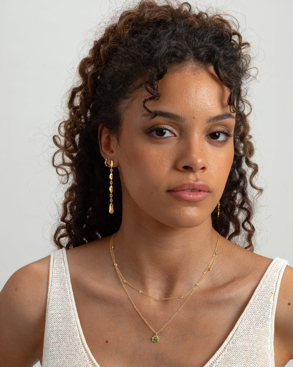 A model styled with Syster P jewelry, wearing two layered gold necklaces—one a simple chain with small discs and the other with a green gemstone pendant—and two different dangling gold earrings.