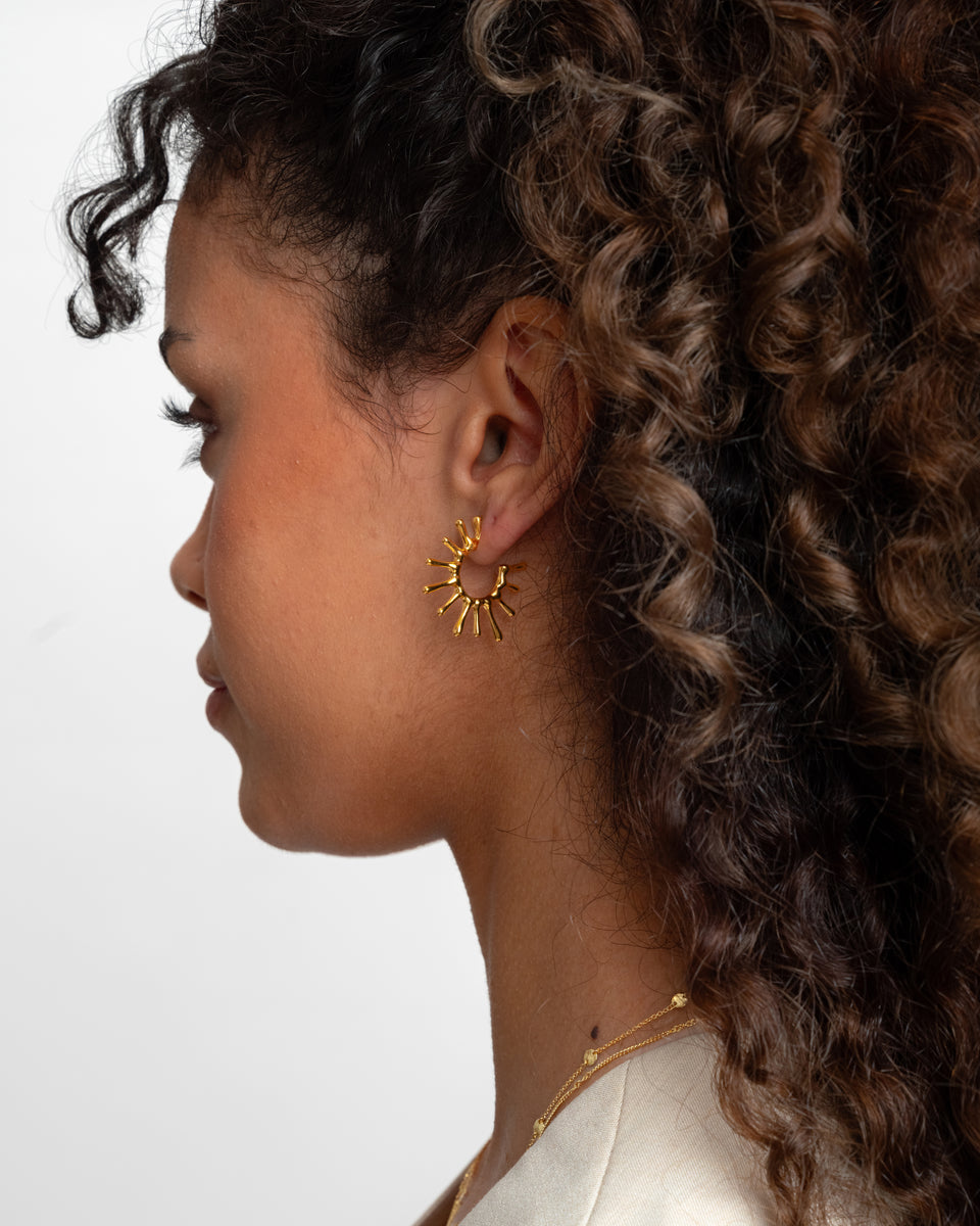 A close-up of a model in profile wearing the Florentina Big Earrings in gold, featuring a unique sun-inspired design, and a delicate gold necklace.