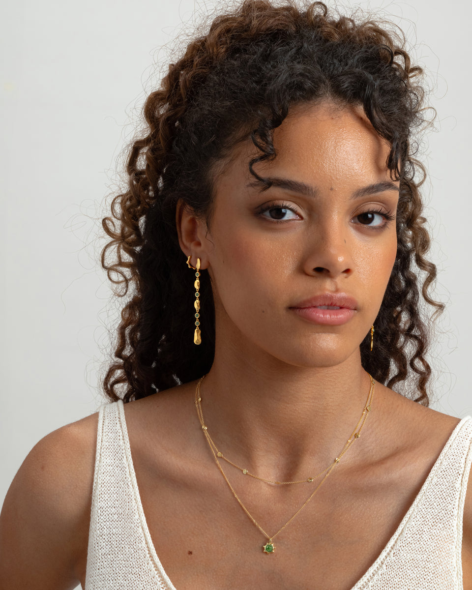 A model wearing the Florentina Long Earrings in gold with green jade, which feature a long dangling design with hammered plates and stones, paired with two layered gold necklaces.