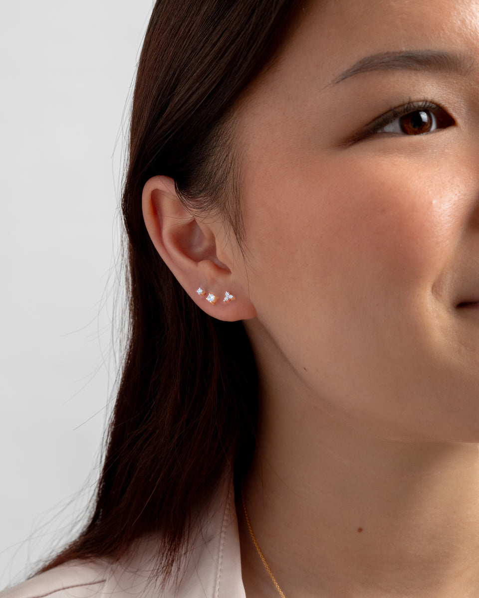 A close-up shot of a model wearing three different gold stud earrings from the Celestia collection, including the Celestia Mini Studs, stacked on her earlobe.