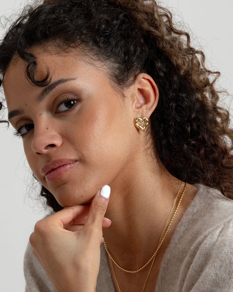 A close-up shot of a model wearing the gold Puff Heart Earrings featuring a chunky and organic heart shape, paired with two layered gold chain necklaces.